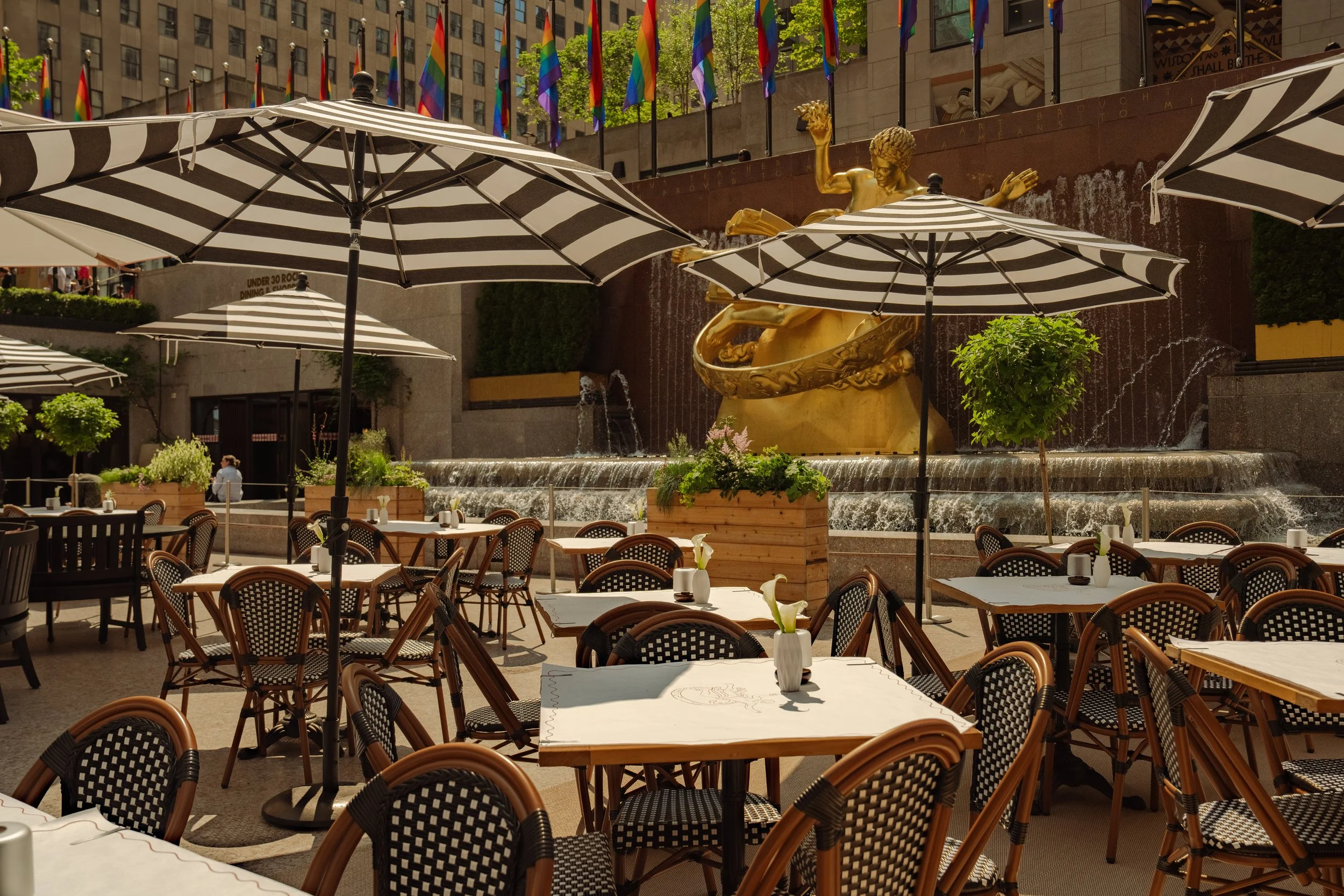 Outdoor cafe with tables and chairs under black and white striped umbrellas in front of a fountain with a gold statue, surrounded by trees and urban buildings, decorated with rainbow flags.