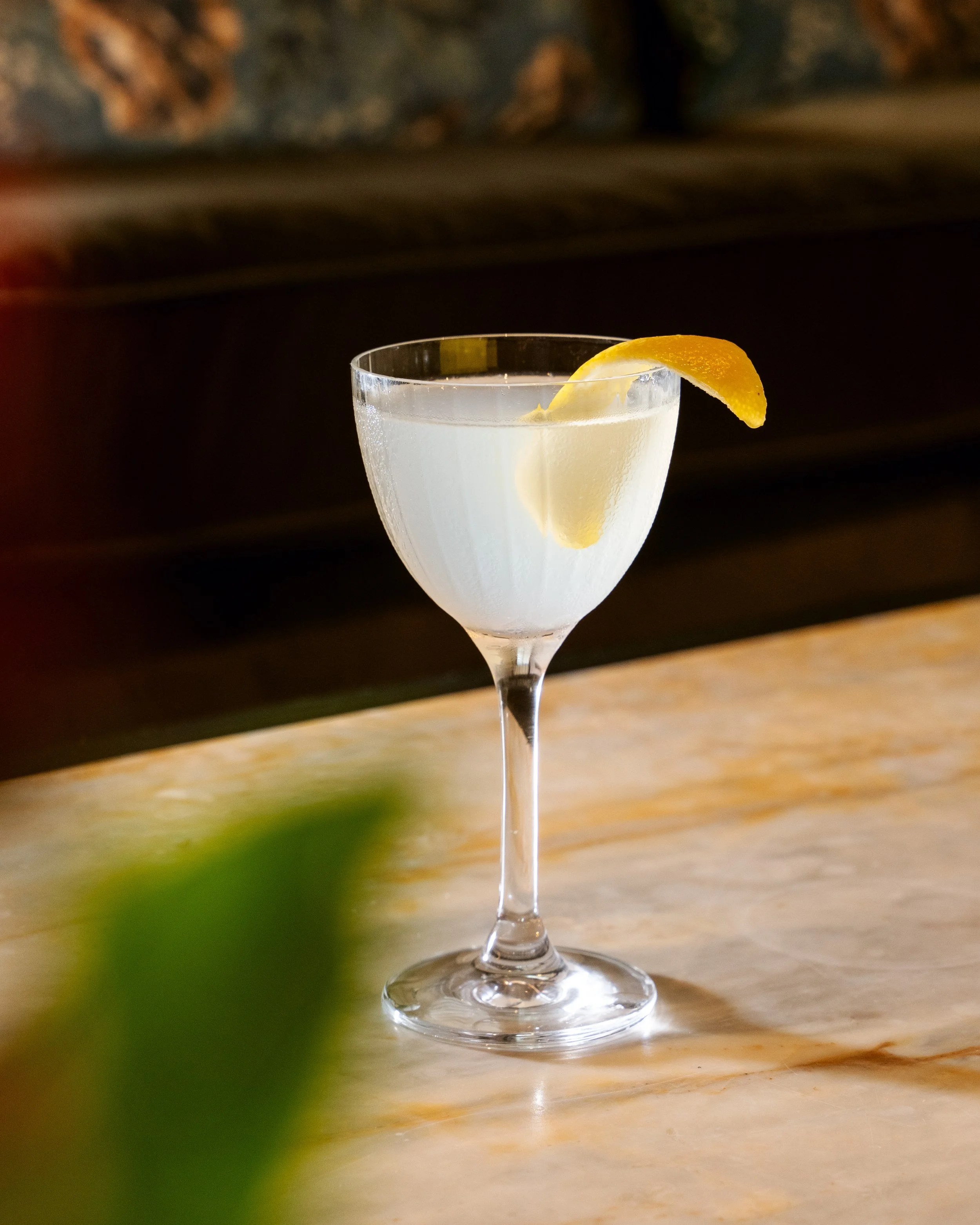 A cocktail glass filled with a clear beverage, garnished with a lemon wedge, on a wooden surface with a dark, blurry background.