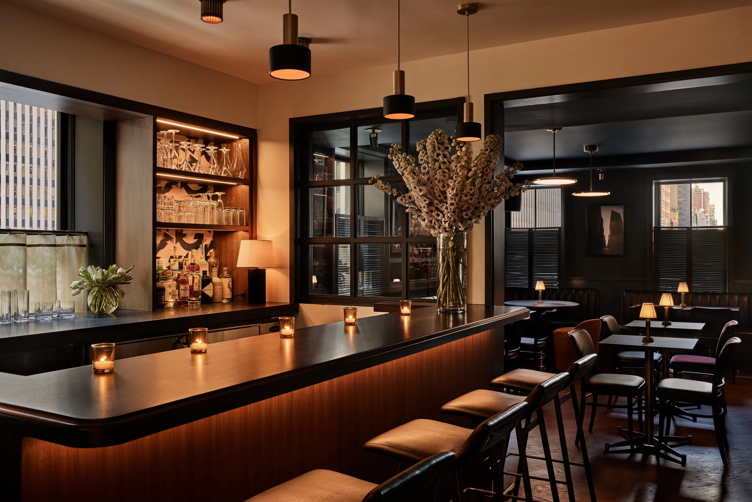 Interior of a stylish bar or restaurant with a long counter, bar stools, a vase with white flowers, a wine and glass display shelf, and warm lighting.