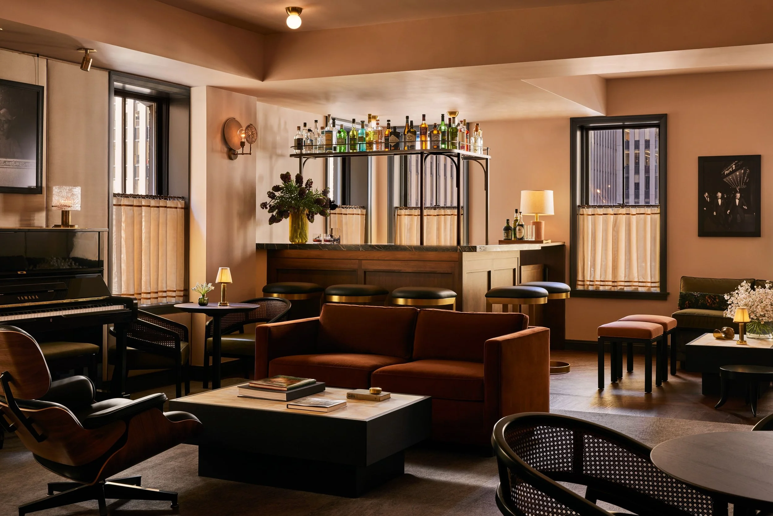 Interior of a stylish bar or lounge with wooden furnishings, a bar counter with a variety of liquor bottles, large windows with curtains, and cozy lighting.