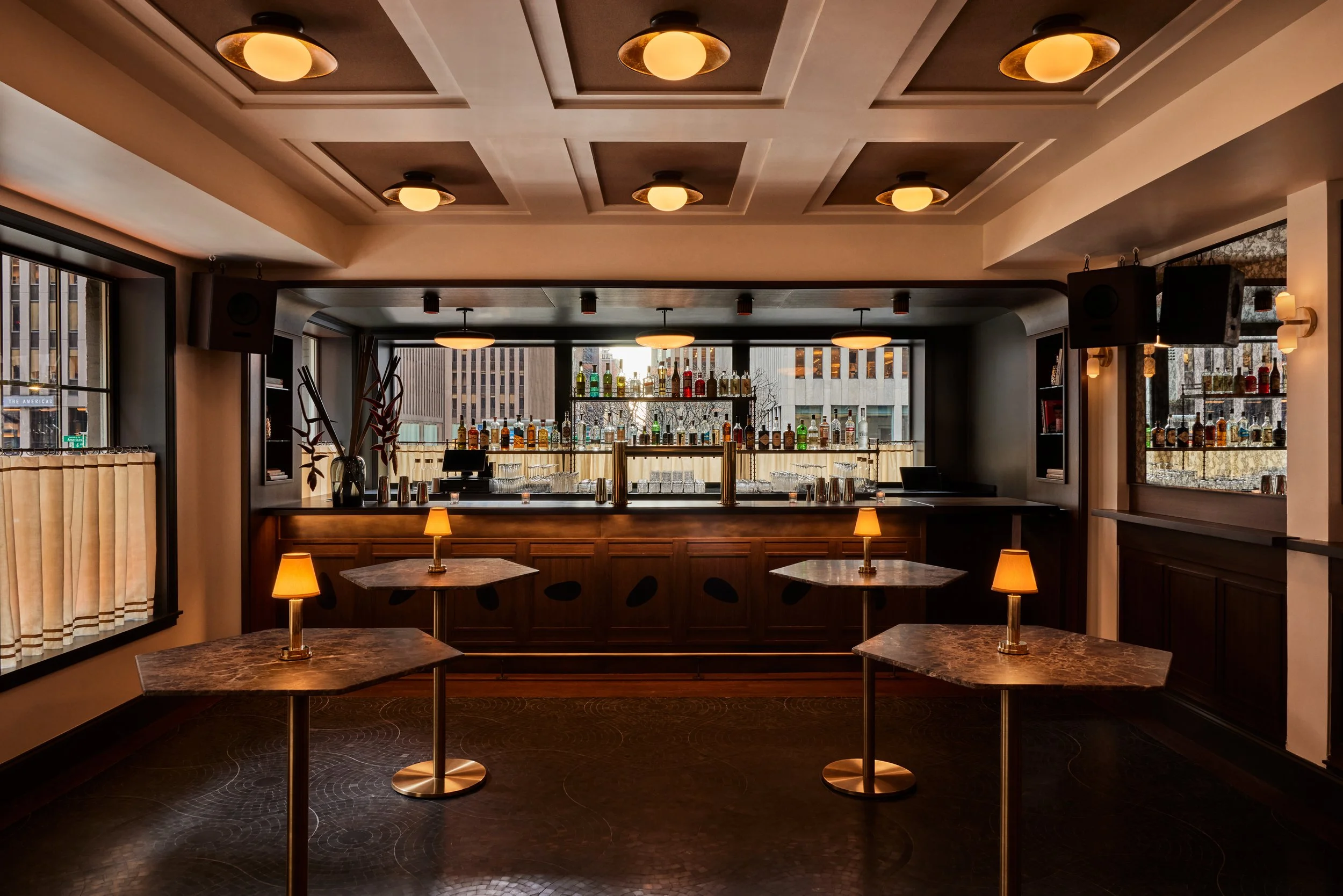 Interior of a modern bar with a marble-top counter, shelves of liquor bottles, and warm lighting fixtures on the ceiling and tables.