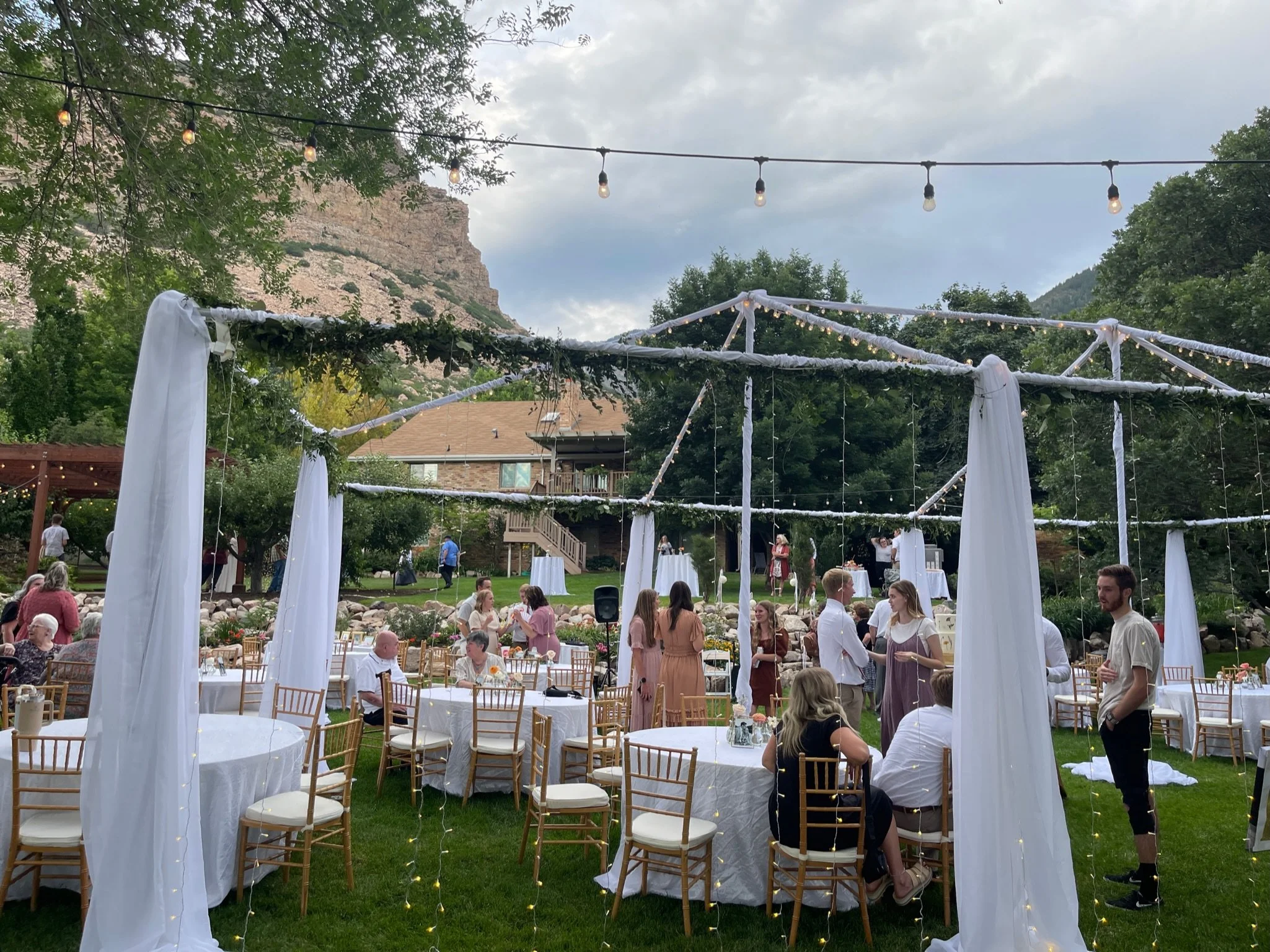 Outdoor wedding reception with round tables, gold chairs, white tablecloths, string lights, and a decorated framework in a garden setting with trees and mountains in the background.
