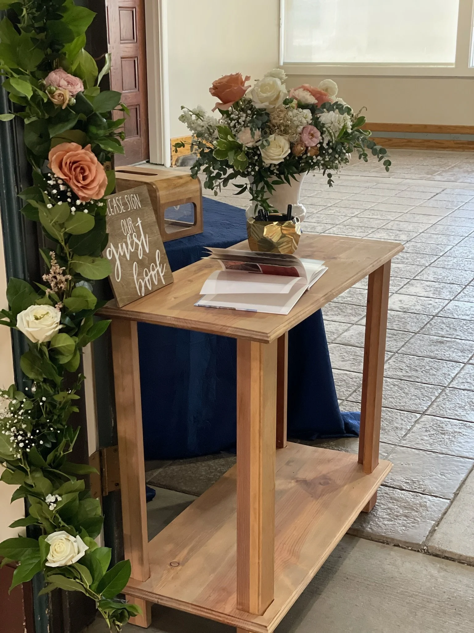 A welcome table decorated with a bouquet of mixed flowers in a white vase, a gold decorative container with pens, a sign that reads 'Please sign our guest book,' and an open guest book on a wooden table, with floral arrangements and greenery around i