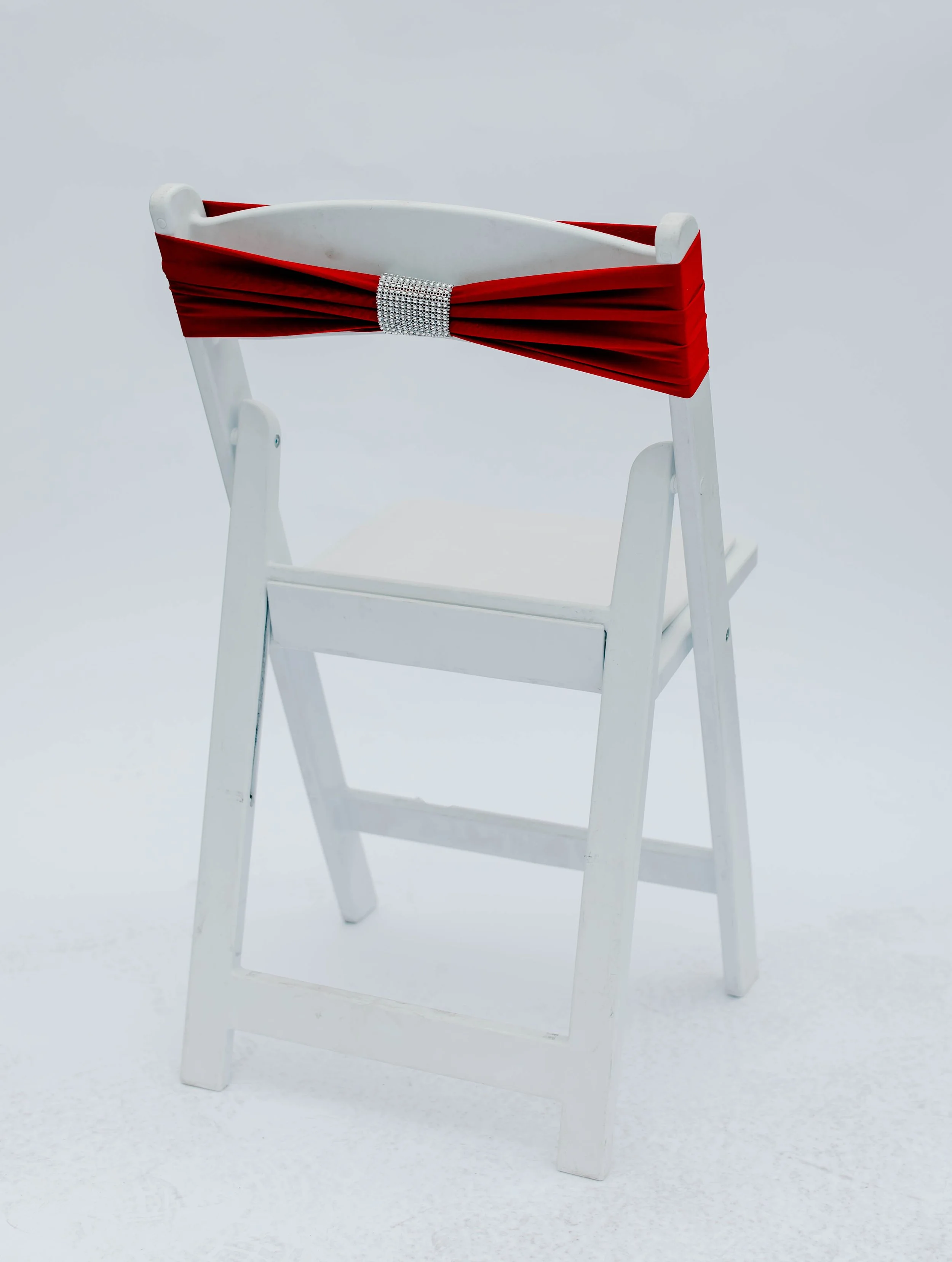 White wooden chair with a red pleated fabric strap and a rhinestone buckle on the backrest, against a plain white background.