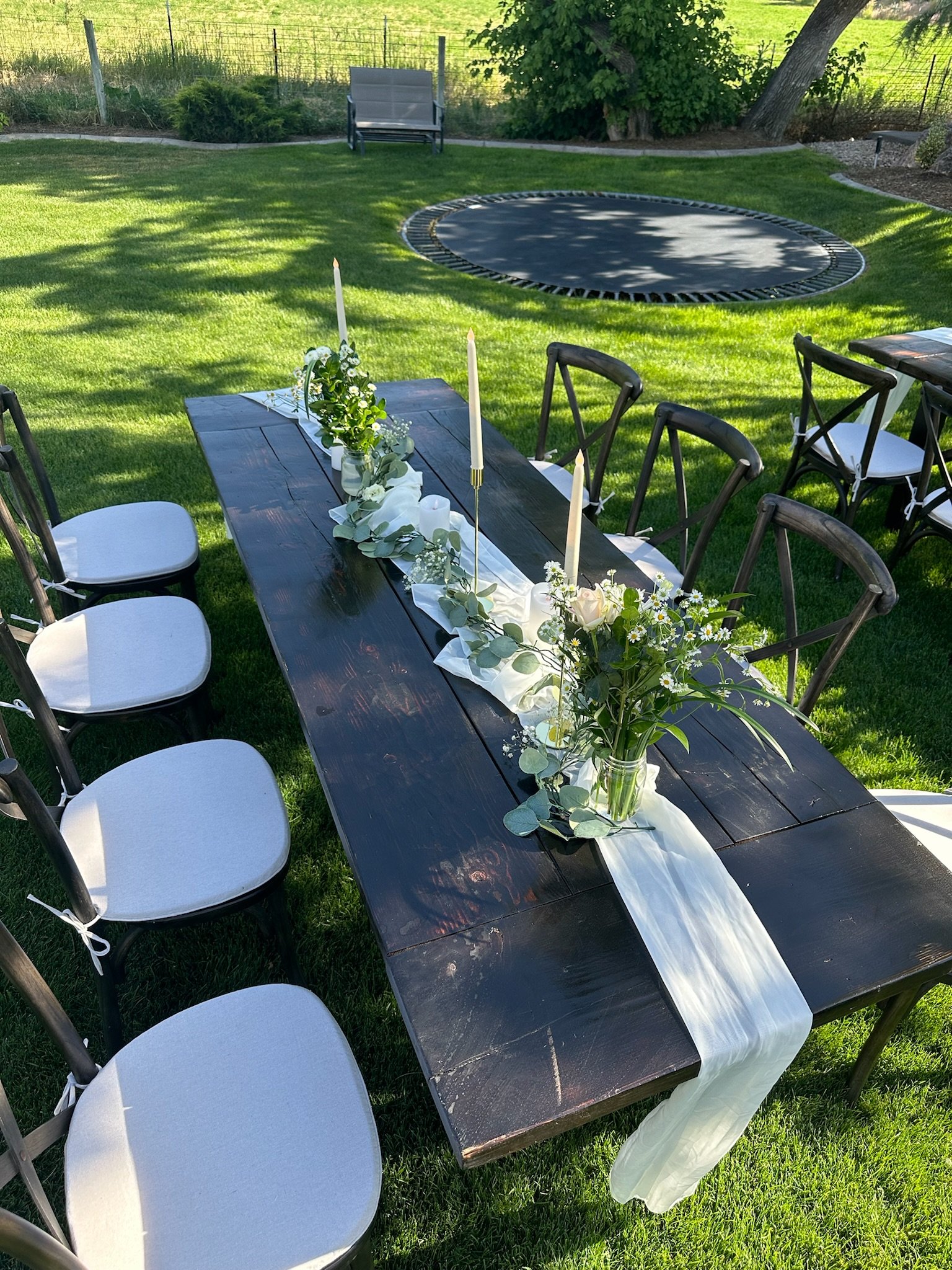 Outdoor dining table with white candles and floral arrangements, set on grass in a backyard with a trampoline and swing in the background.
