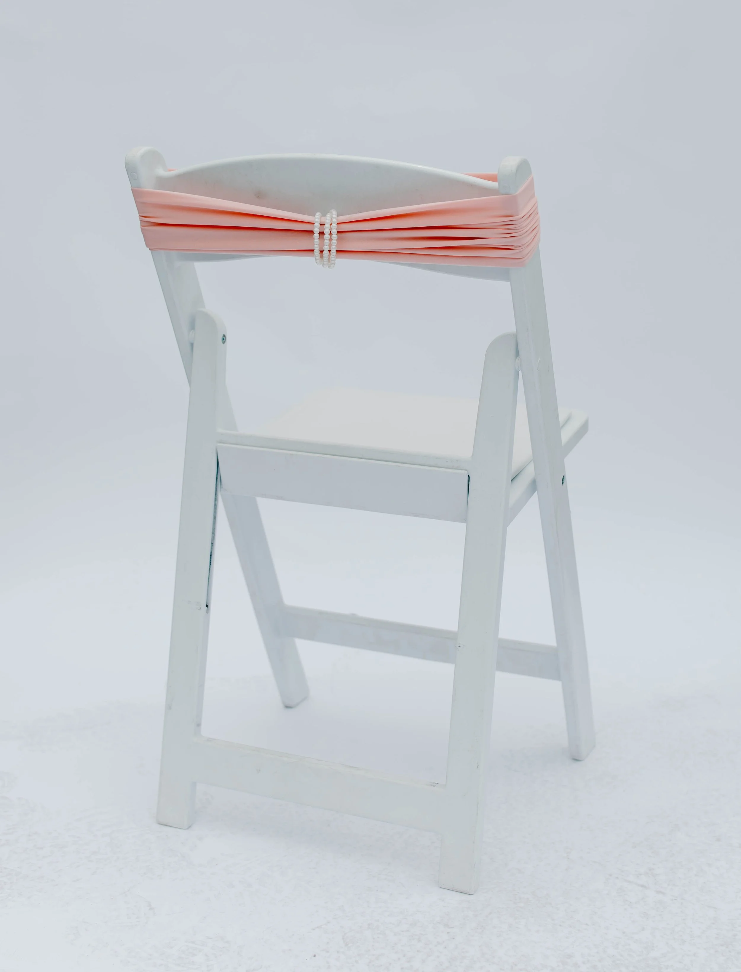 White wooden high chair with pink fabric draped over the backrest, held with a string of white beads, against a plain light background.