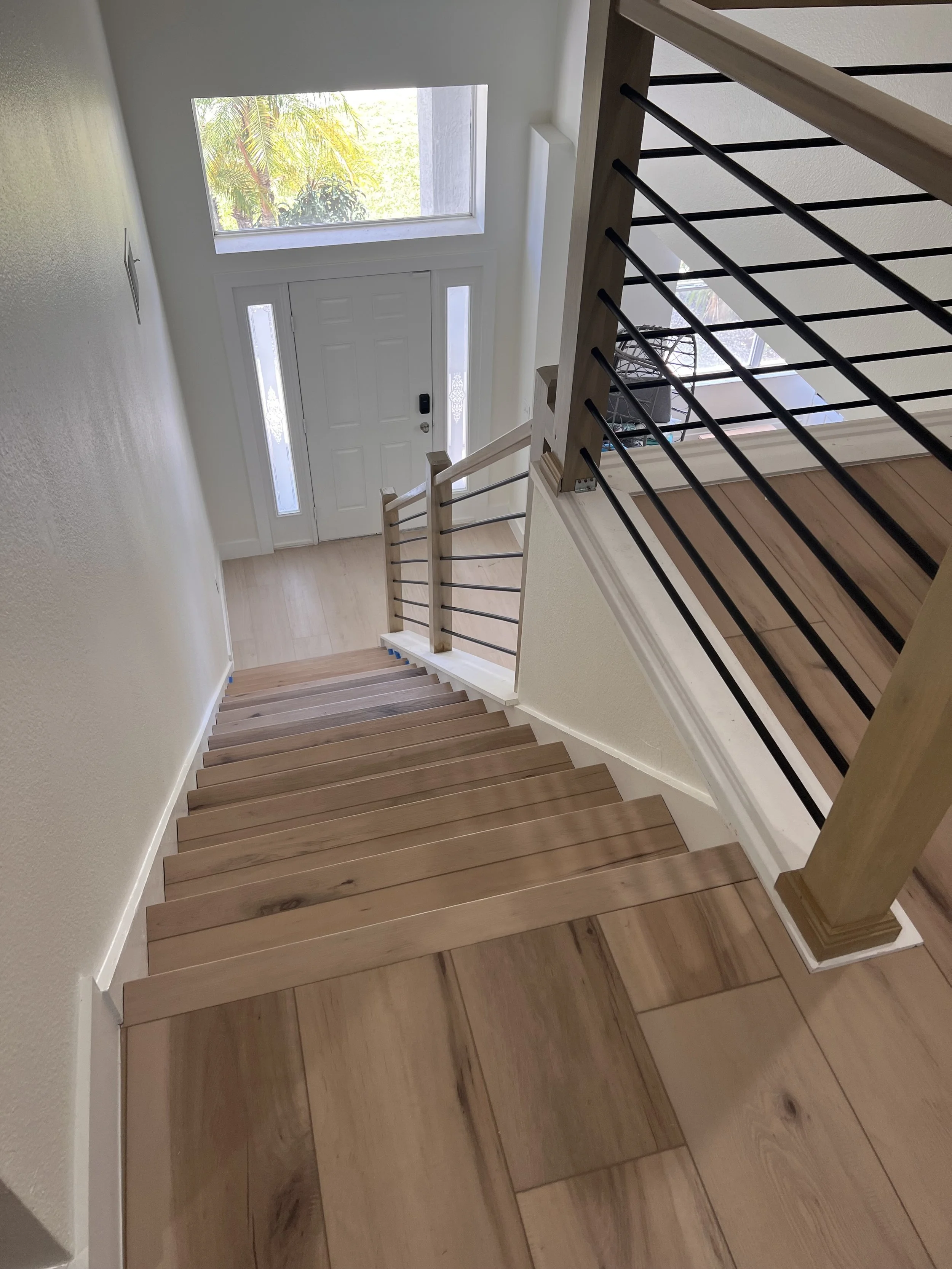 Interior view of a staircase looking towards the front door with a large window, wooden stairs, and black railing.