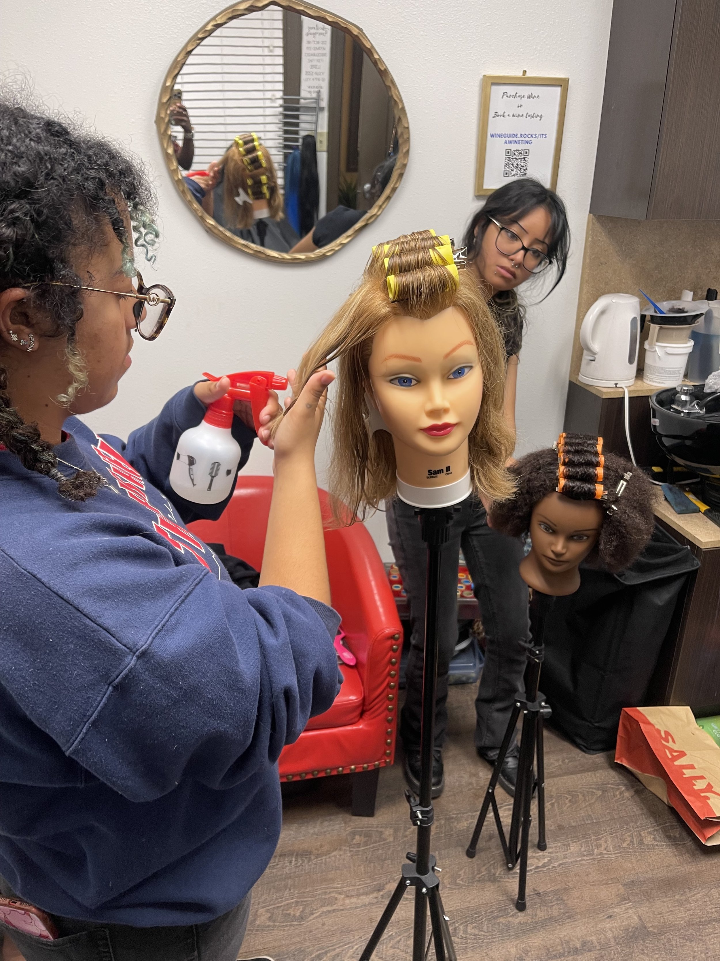 A hairstyling class in progress with a hair mannequin on a stand, students assisting and observing.