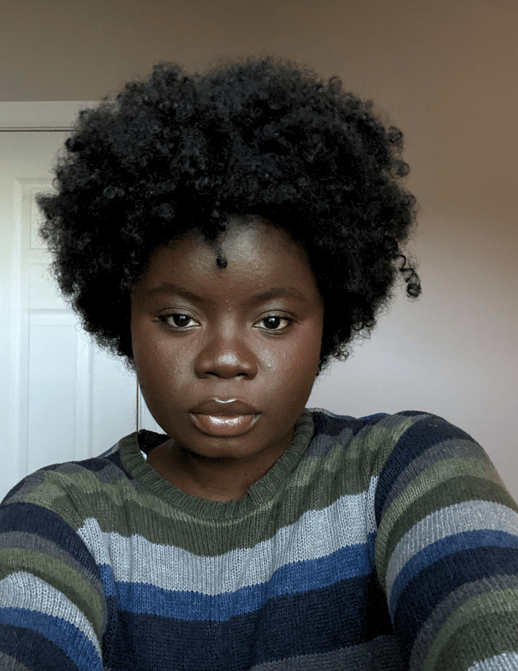 A young woman with dark skin, short curly hair, and wearing a multicolored striped sweater taking a selfie in front of a plain background.