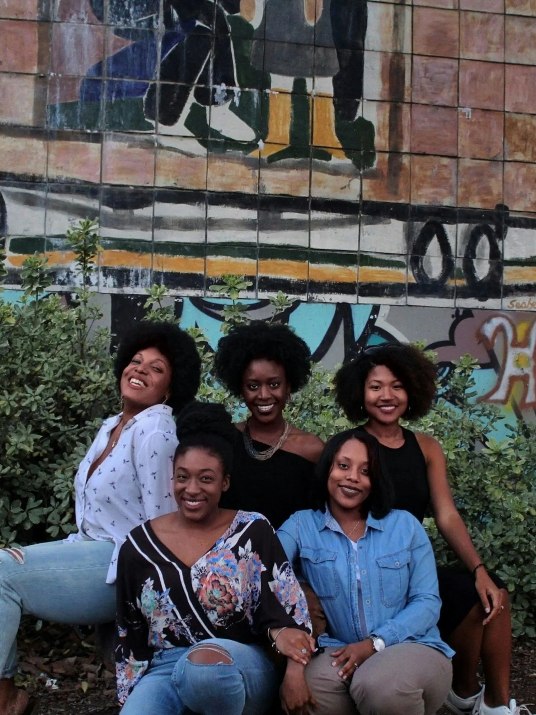 Five women posing outdoors, with a mural painted on a brick wall behind them and green bushes in front.