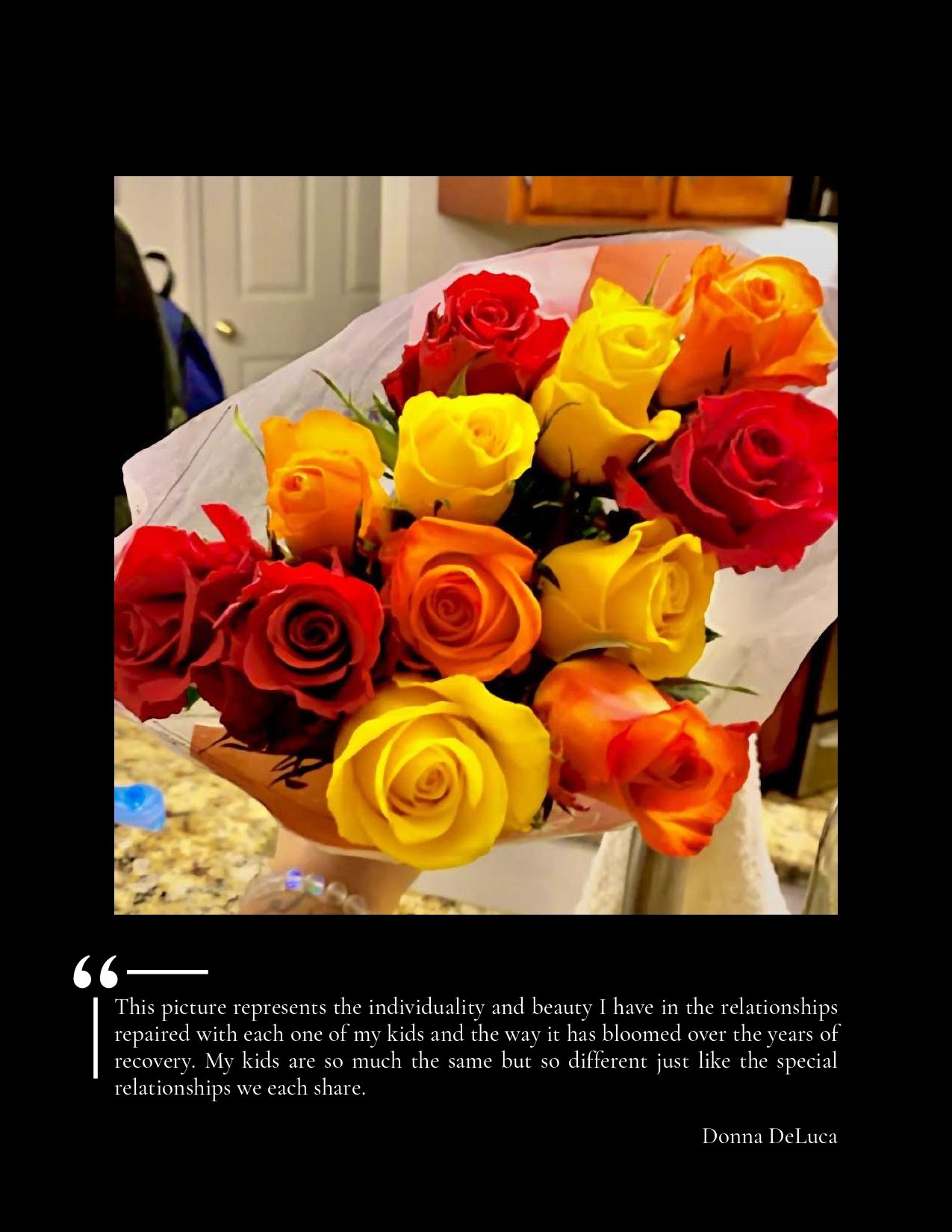 A bouquet of multicolored roses including red, yellow, orange, and peach roses, wrapped in white paper, held against a background featuring a kitchen countertop and a door.