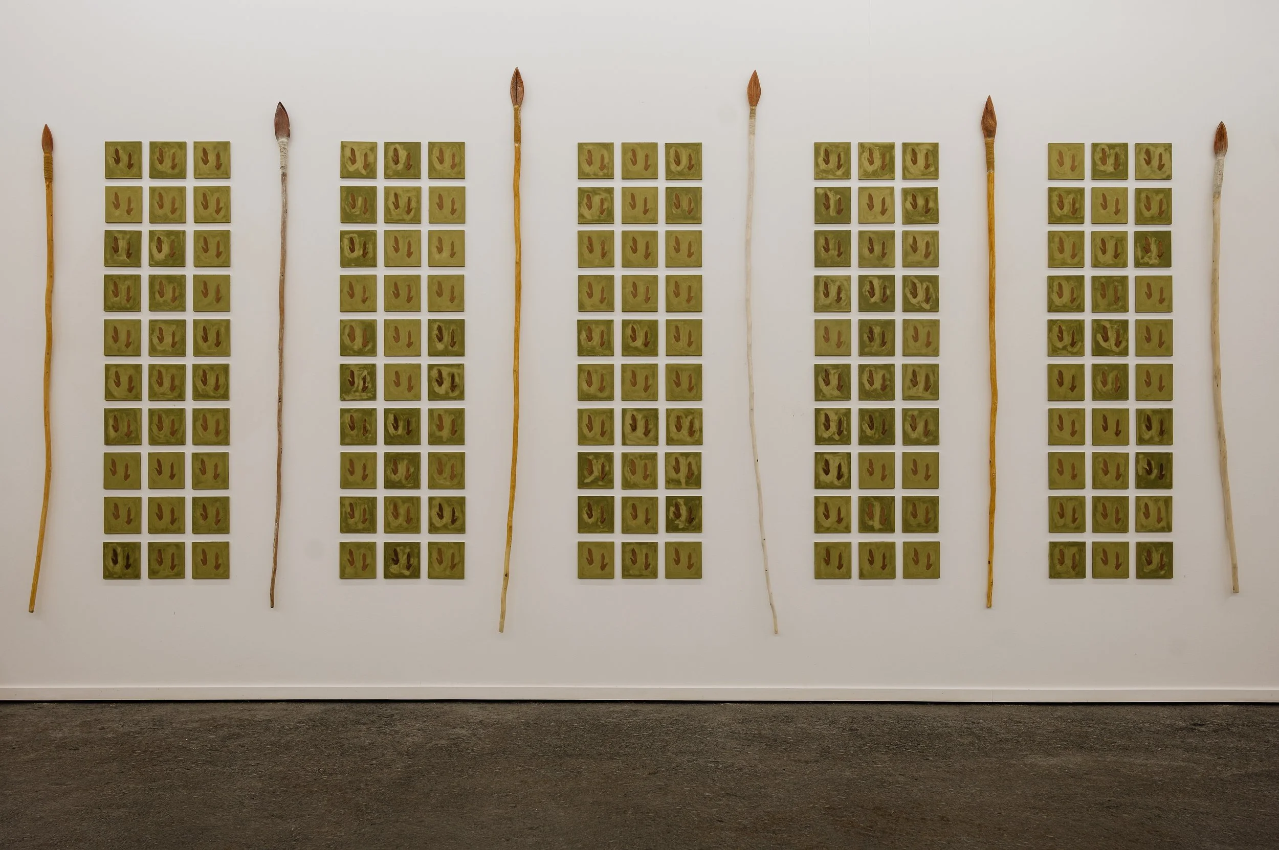 Art installation featuring six vertical rows of small framed images arranged in columns, with long wooden sticks placed between the column groups on a plain white wall.