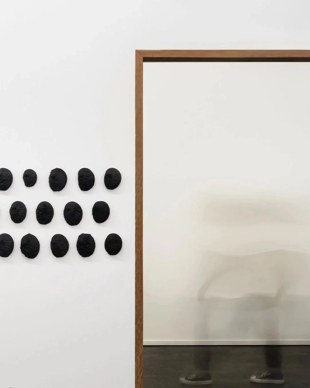 An art installation featuring a white wall with 16 black textured oval objects arranged in a grid, and a partial wooden frame to the right with a reflection of a person walking.