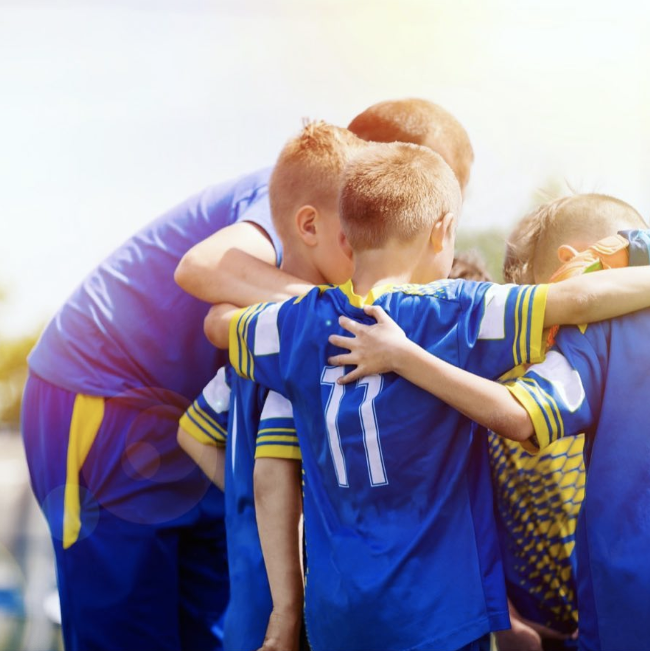Youth soccer team huddle with their coach outside on a sunny day.