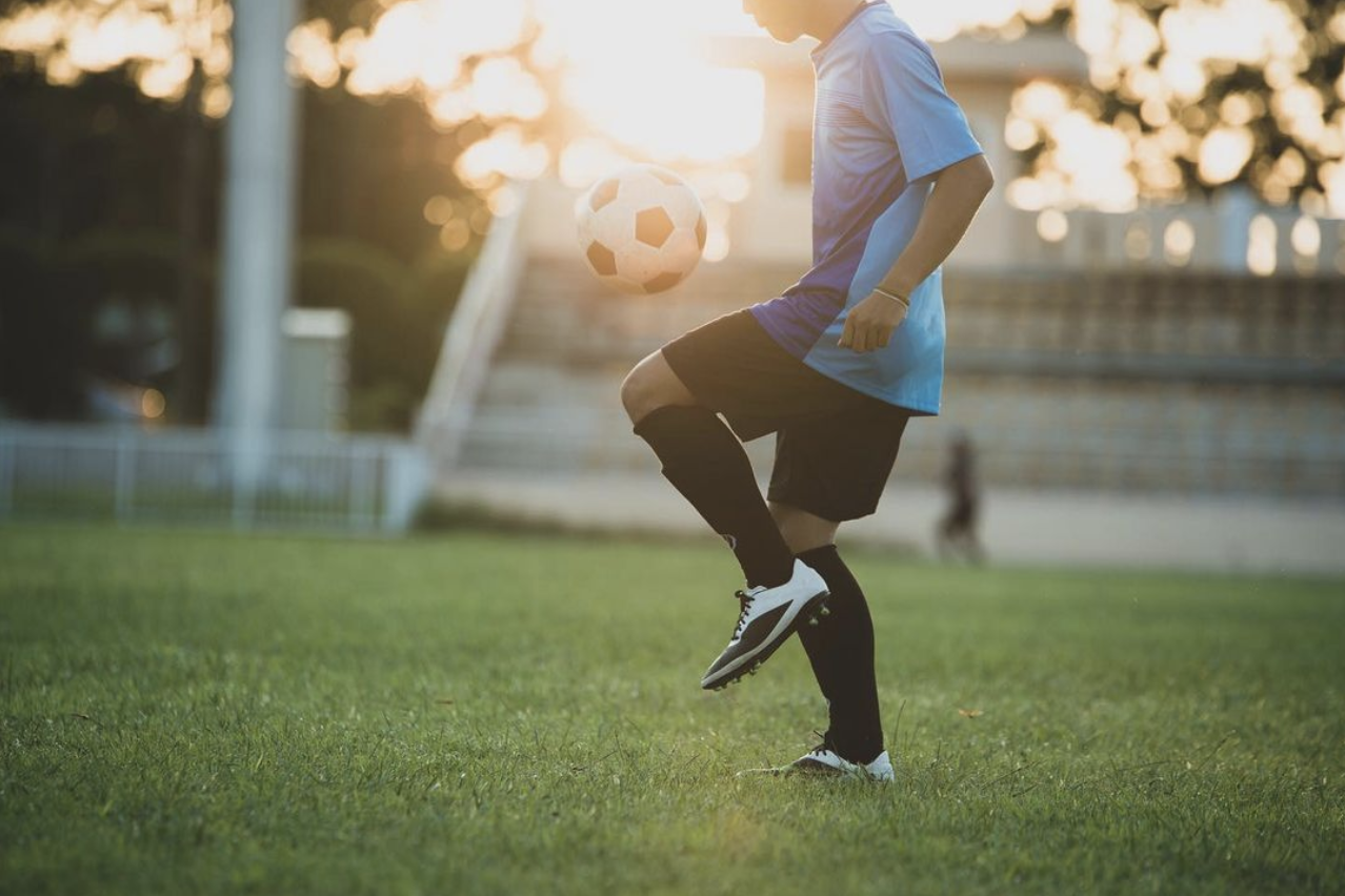 A soccer player in a blue jersey and black shorts practicing on a field at sunset, doing a juggling move with a soccer ball.