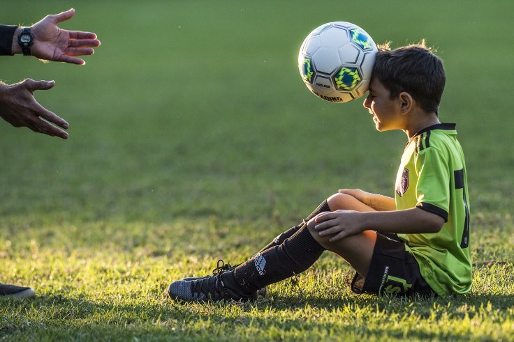 A young boy in a yellow soccer uniform sitting on grass with a soccer ball balancing on his head, while two people reach out towards him.