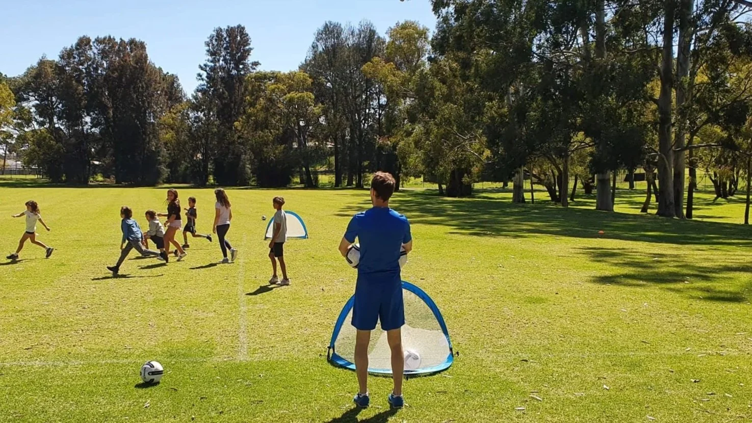 Children and adults playing soccer on a grassy field in a park with trees in the background on a sunny day.