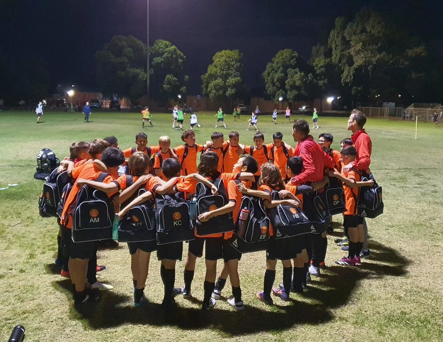 A youth soccer team wearing orange jerseys and black shorts gather in a circle on a field at night, hugging each other. Two coaches in red shirts are with the team. In the background, other players are playing soccer on the field under bright stadium