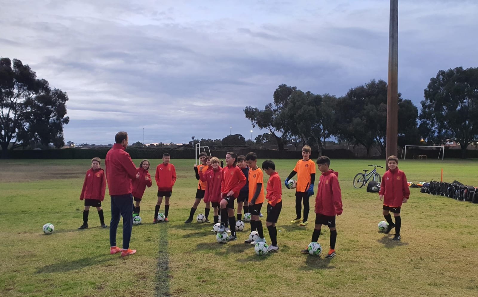 Group of young soccer players with coach on a grassy field during dusk, some holding soccer balls, with trees and goal posts in the background.