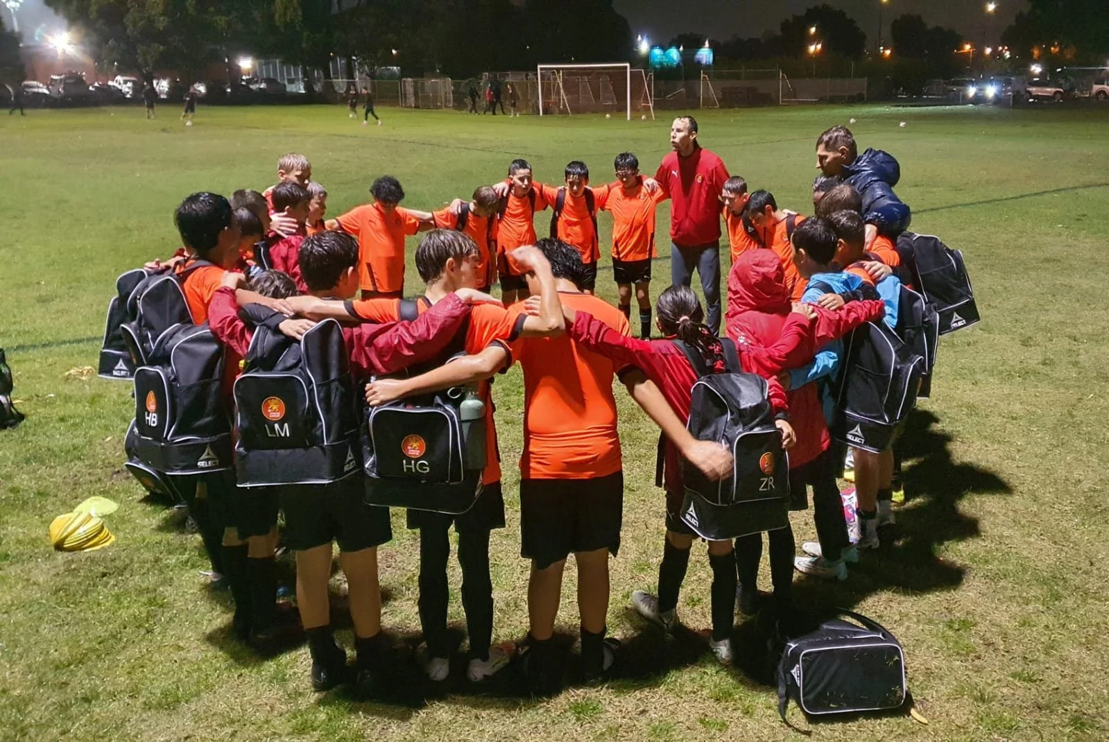 A group of young soccer players in red and orange uniforms and their coaches huddle together on a soccer field at night, with grassy area, goalposts, and parked cars in the background.