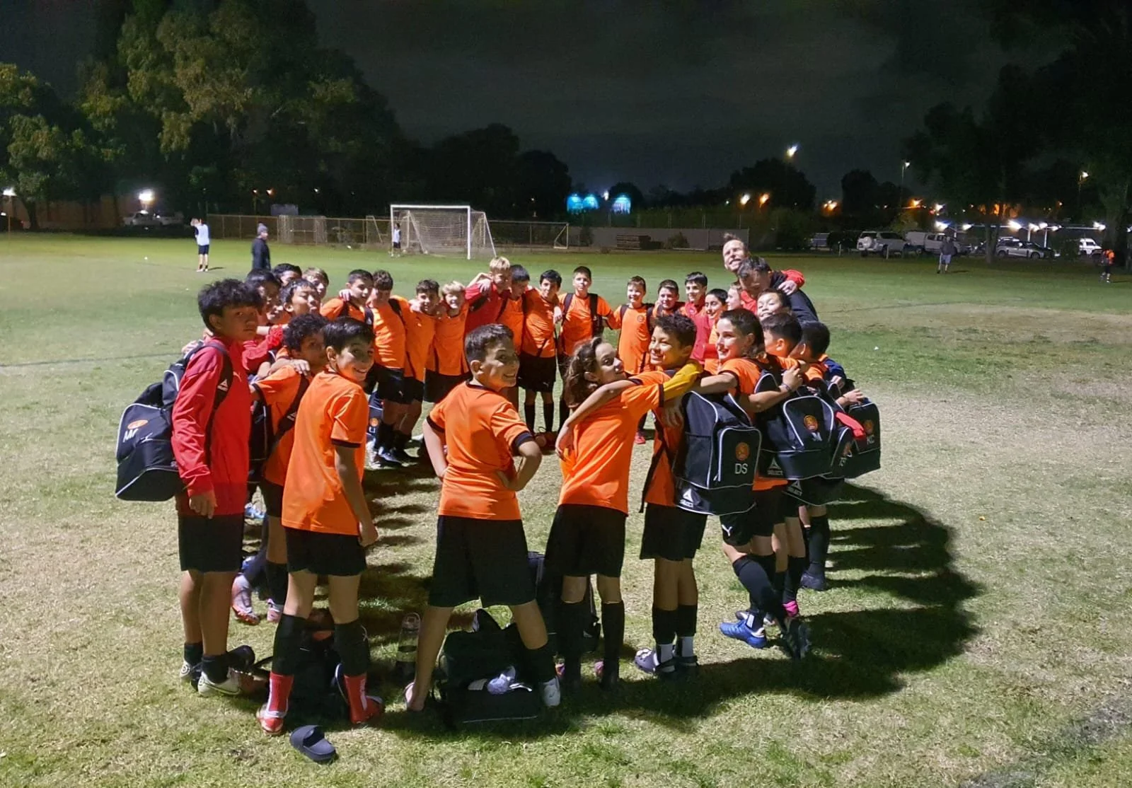 A group of young soccer players in orange jerseys and black shorts stand in a circle on a soccer field at night, smiling and hugging each other, with their coach, also smiling, joining the circle. Some players have backpacks, and others are holding w