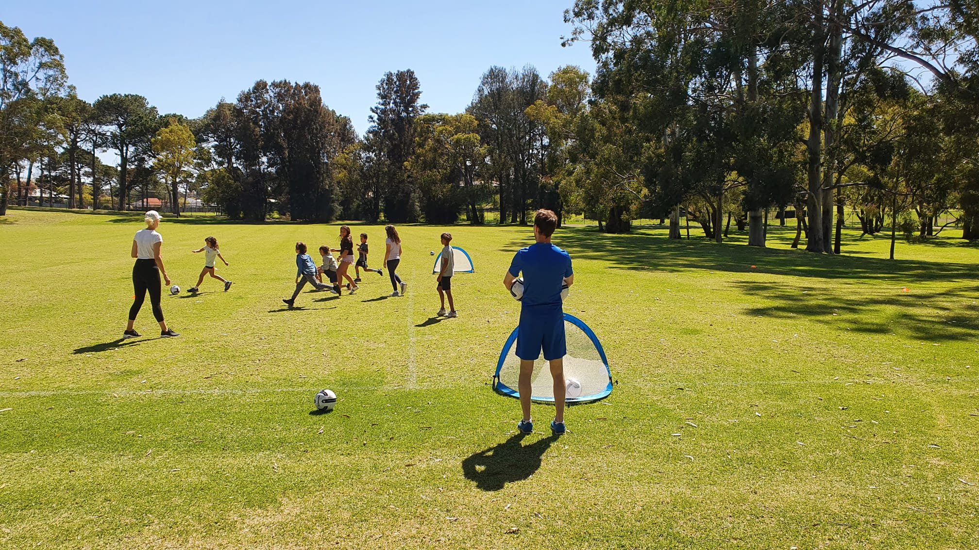 Children and adults playing soccer on a grassy field in a park with trees in the background on a sunny day.