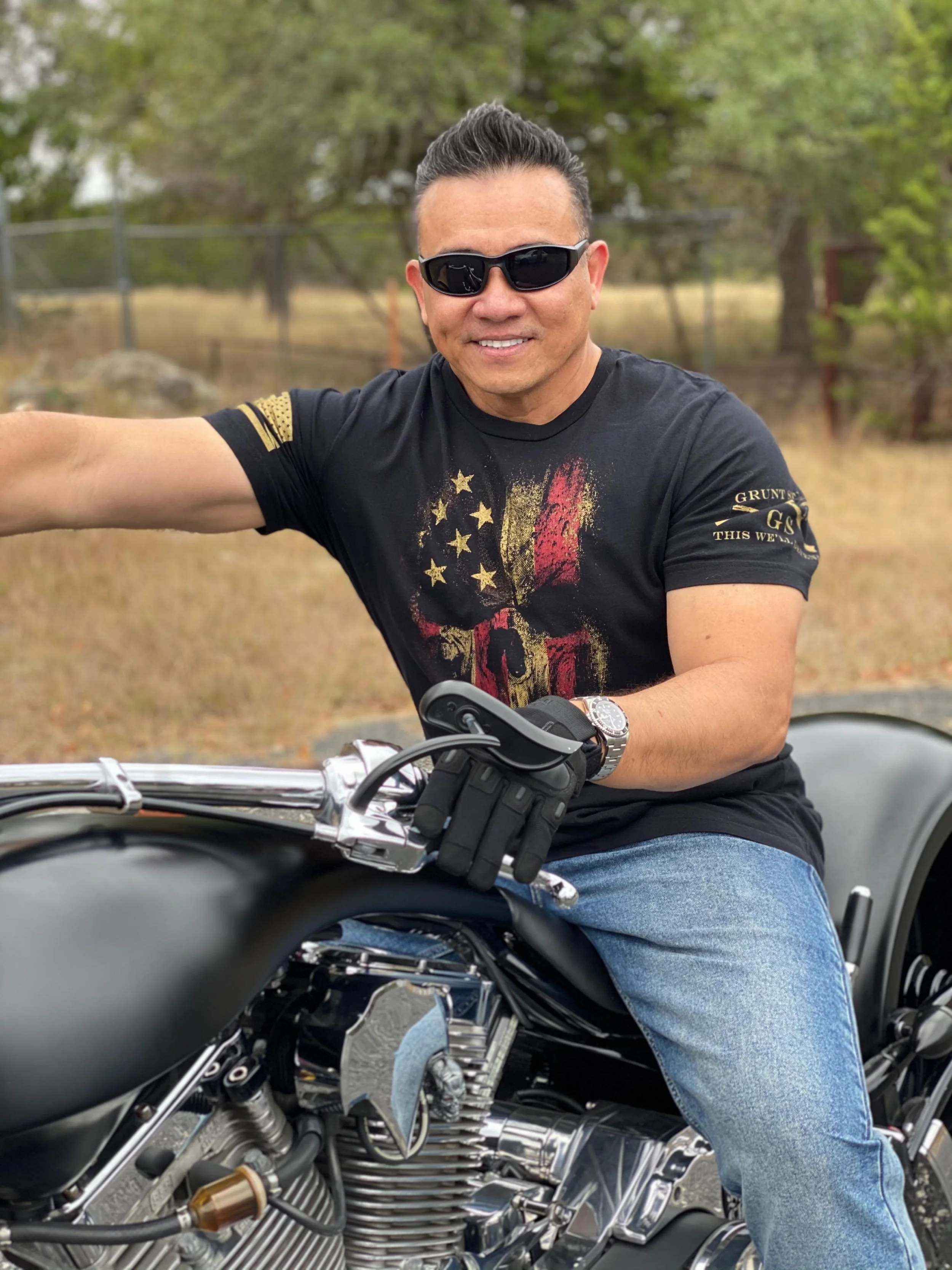 A man wearing sunglasses and a black T-shirt with a graphic design, sitting on a black motorcycle outdoors with trees and grass in the background.