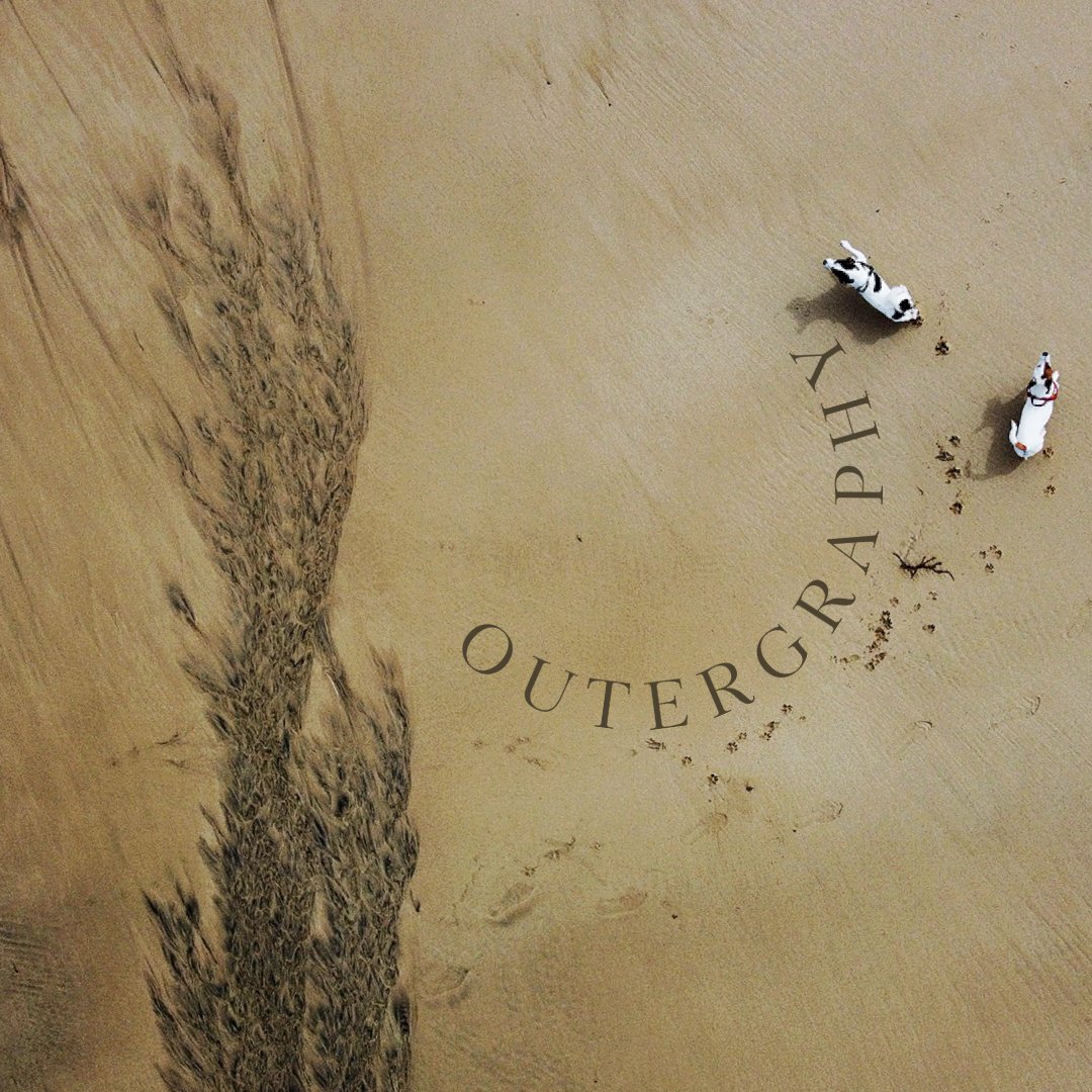 Two dogs walking on a sandy beach near a rock formation, leaving footprints behind, with the logo of Outergraphy written in the sand.