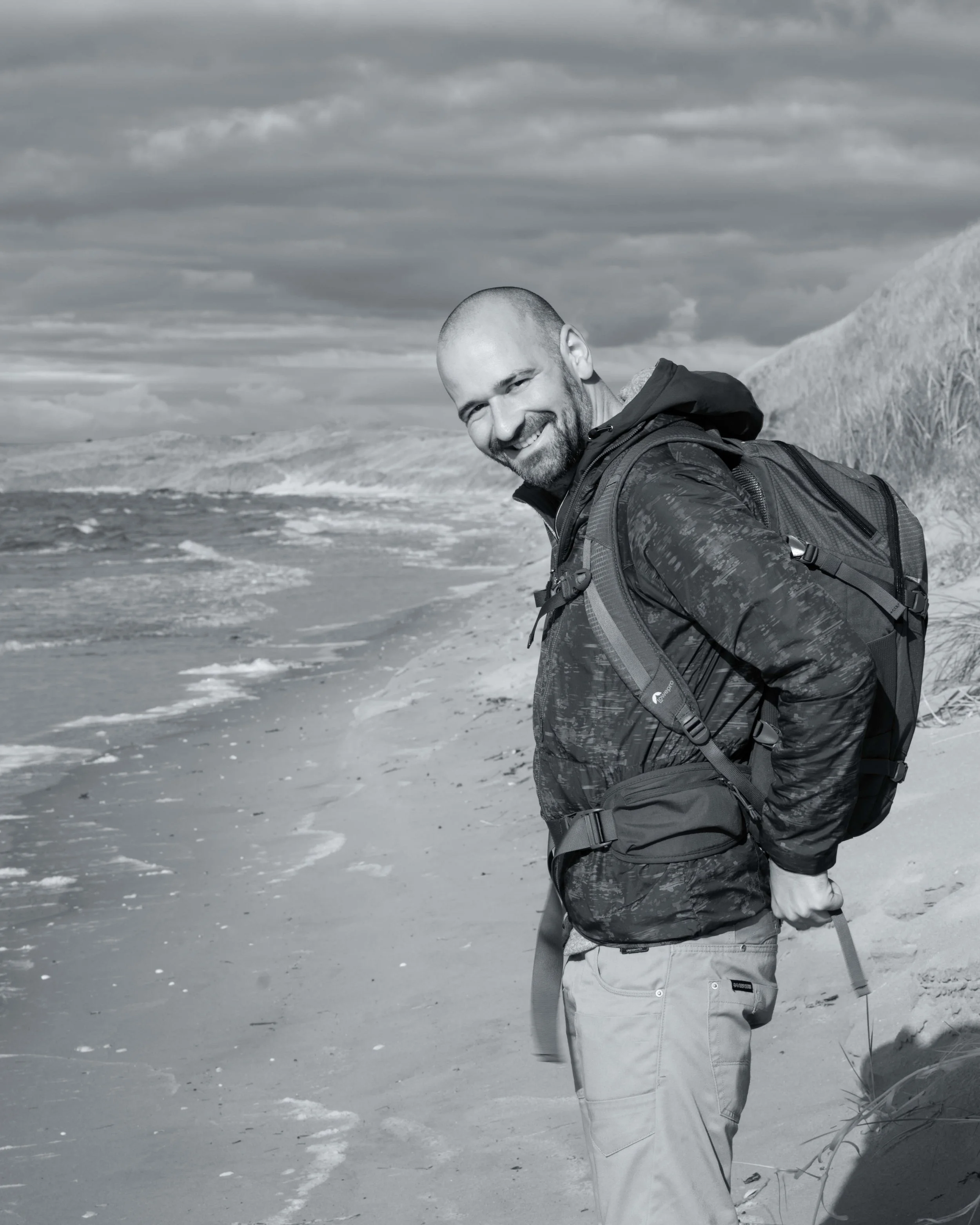 Smiling man on beach wearing a backpack and holding walking poles, with clouded sky and shoreline in the background.