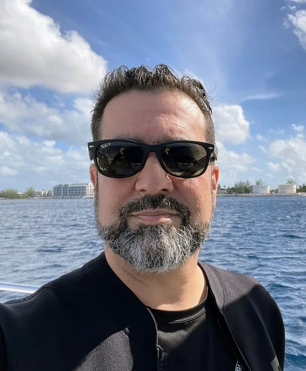 Man wearing sunglasses with a beard, standing near the water with a cityscape and cloudy sky in the background.