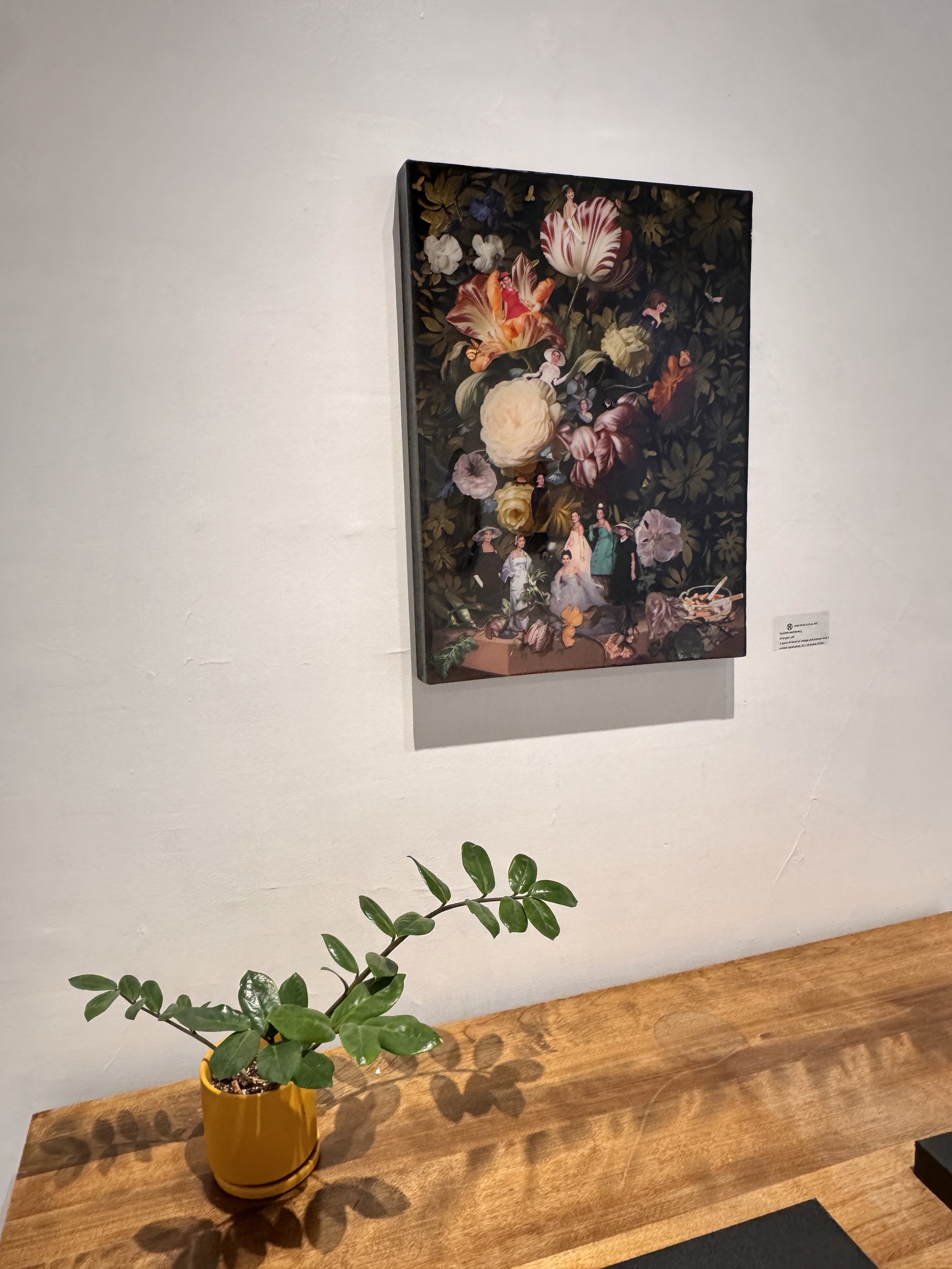 A potted green plant with multiple leaves on a wooden table, with a background painting of flowers and small human figures.