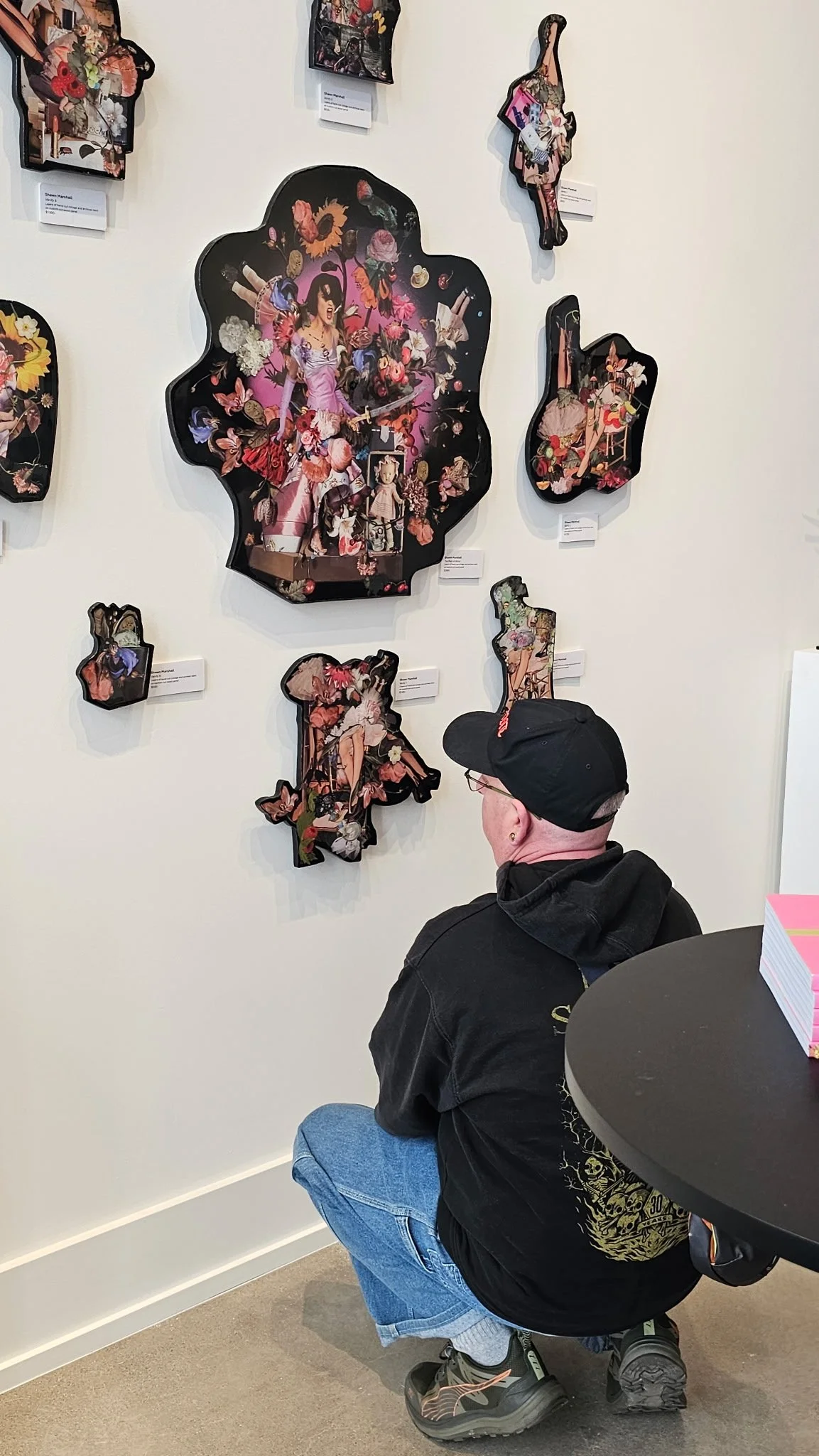 A person kneeling in front of a wall art display at an art gallery. The wall features several black-shaped artworks with colorful floral and pop culture collage designs. The person is wearing a black jacket, blue jeans, a black cap, and sneakers.