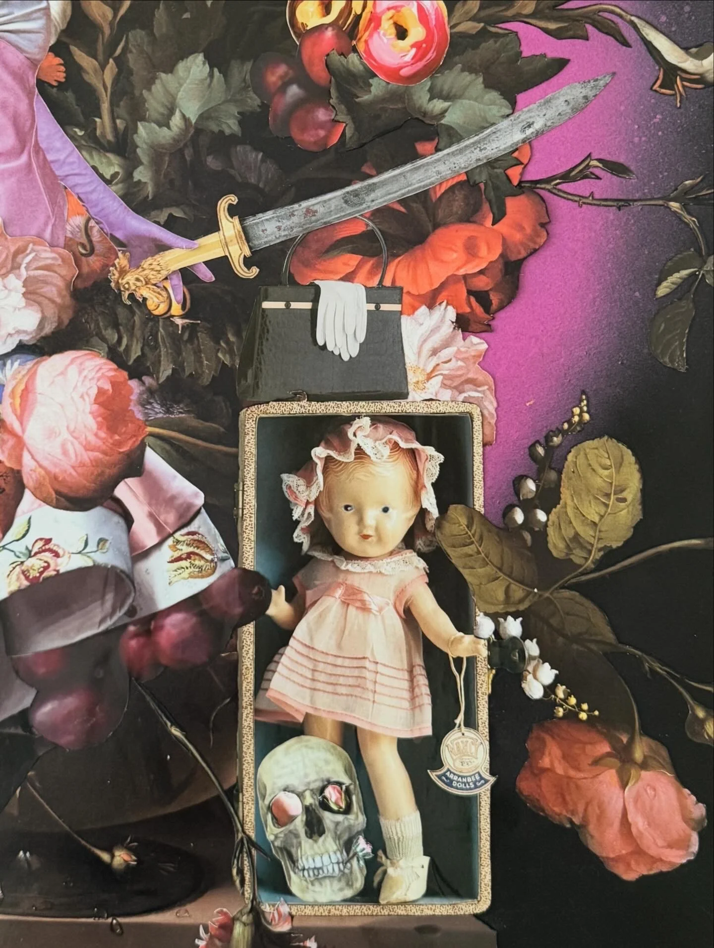 A vintage porcelain doll with blonde hair, wearing a pink dress, lying on a black surface amidst flowers, leaves, and various objects including a skull, a sword, and a purse.