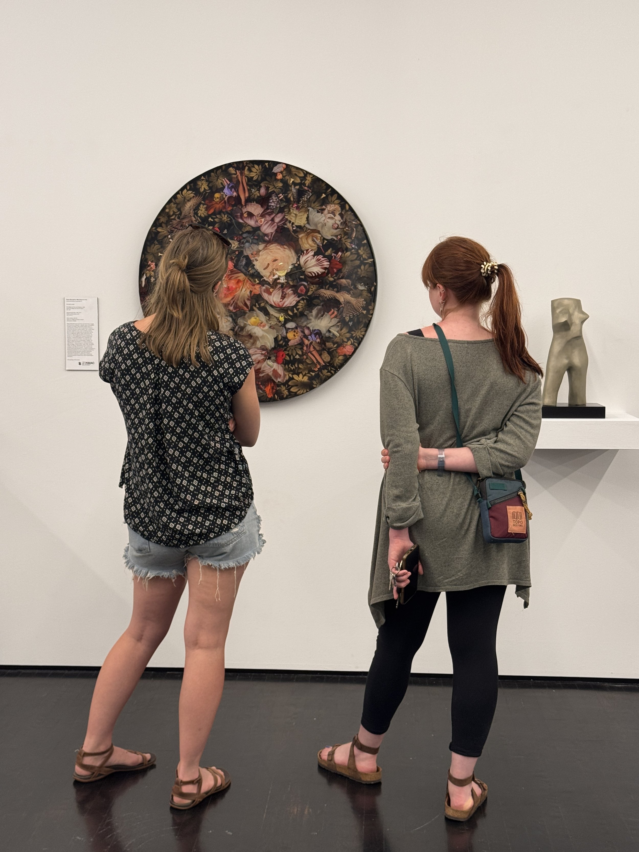 Two women are standing and looking at a large circular artwork with a floral and animal design on a white gallery wall. One woman has blond hair, is wearing a black and white patterned shirt and denim shorts, and sandals. The other has red hair tied 