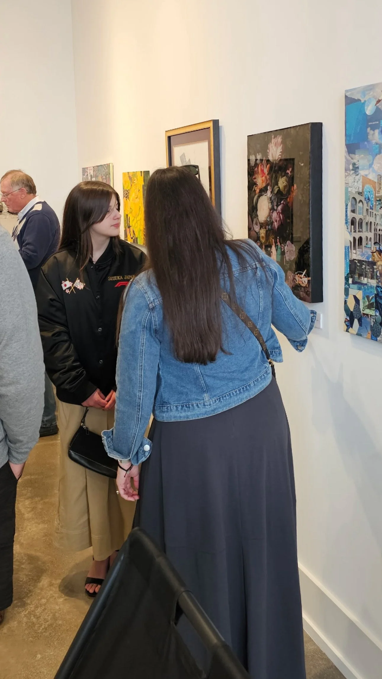 Two women in conversation at an art gallery, with various paintings on the white wall behind them. One woman has long dark hair and is wearing a teal jacket and black skirt, the other has long dark hair and is wearing a black jacket with local patche