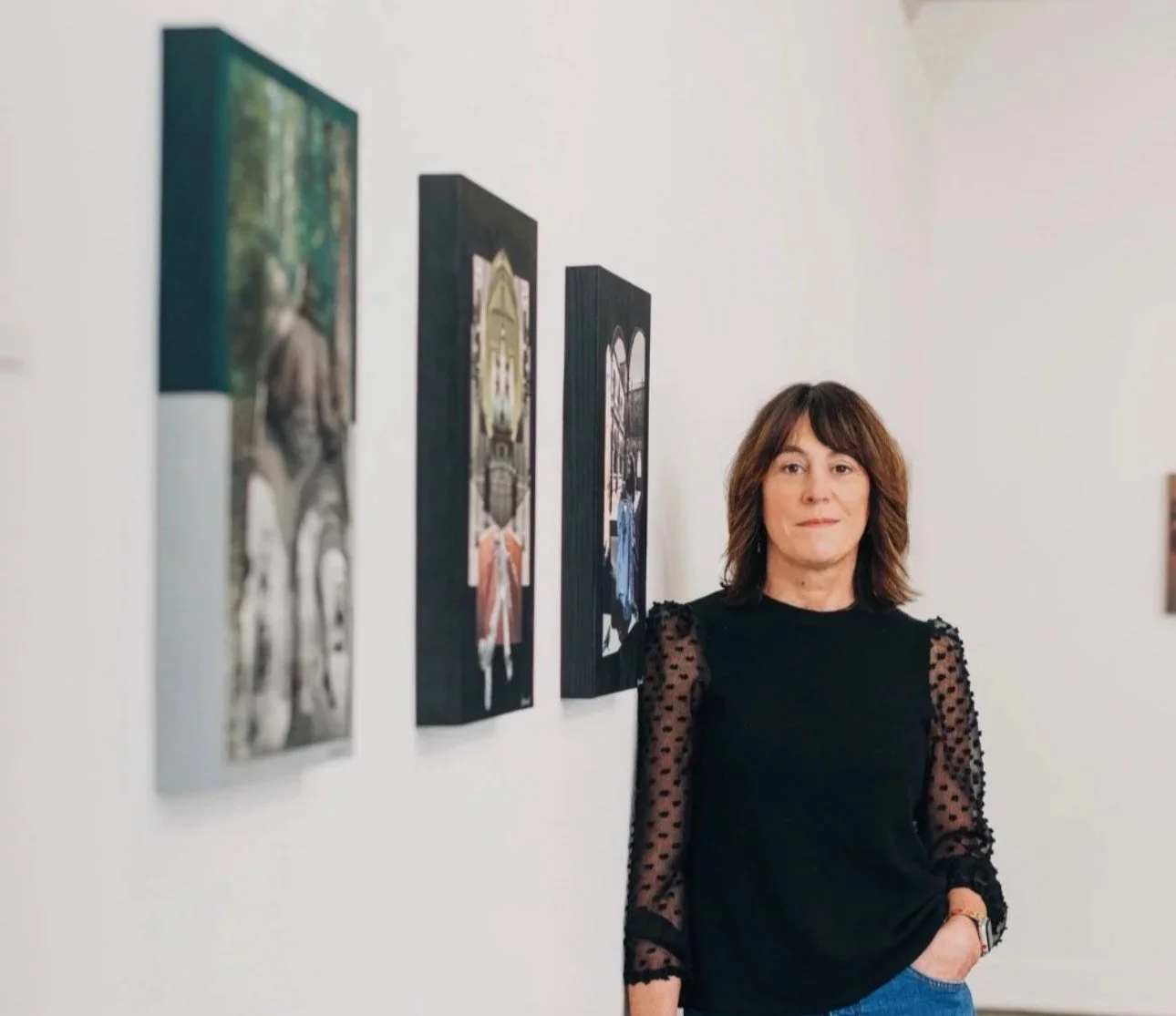 A woman with shoulder-length brown hair standing in an art gallery with white walls, wearing a black top with sheer, polka-dotted sleeves, and looking at the camera. There are four colorful artworks hanging on the wall beside her.
