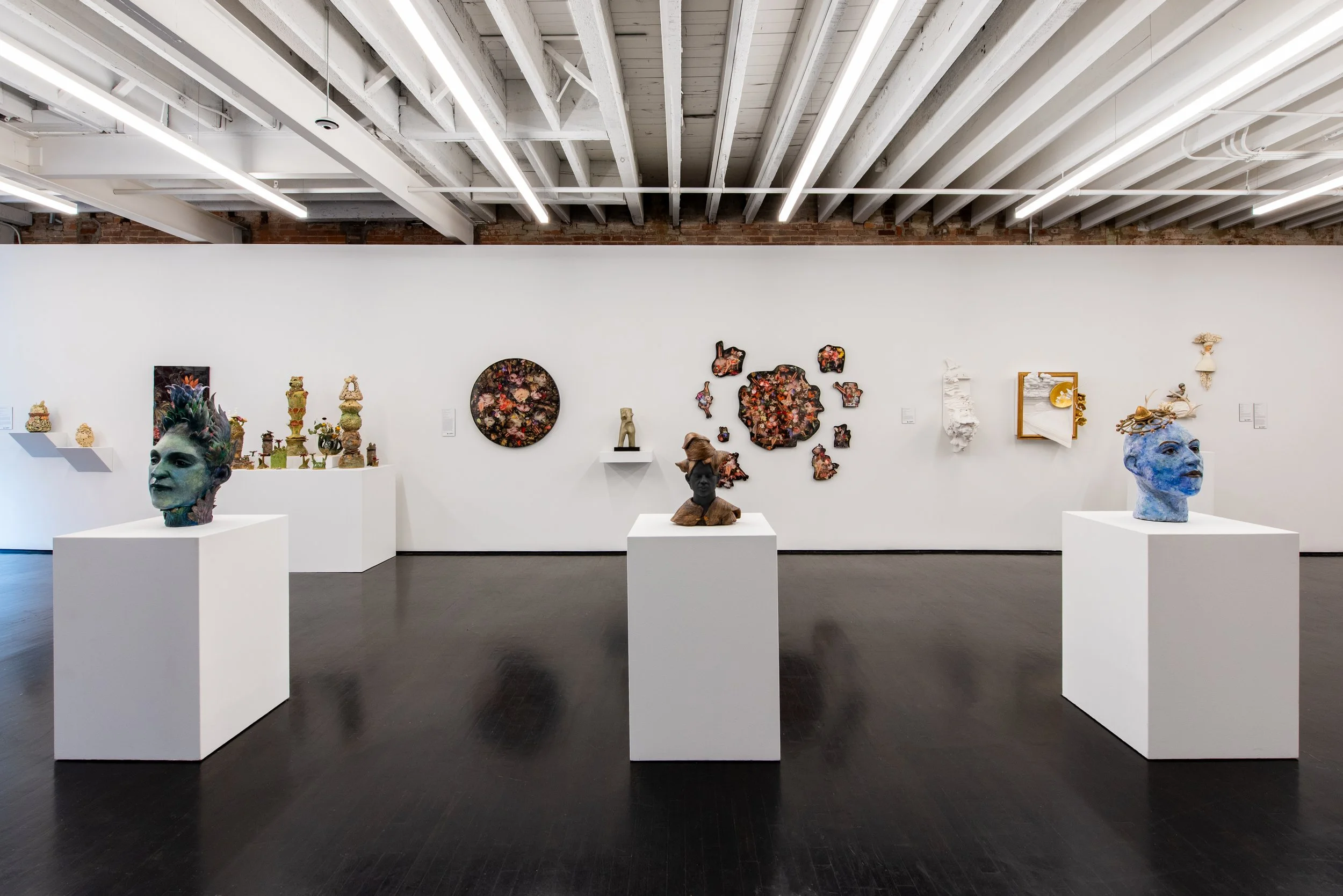 An art gallery displaying various sculptures, including busts, decorative pieces, and abstract works, all arranged on white pedestals against a white wall with wall-mounted art pieces.