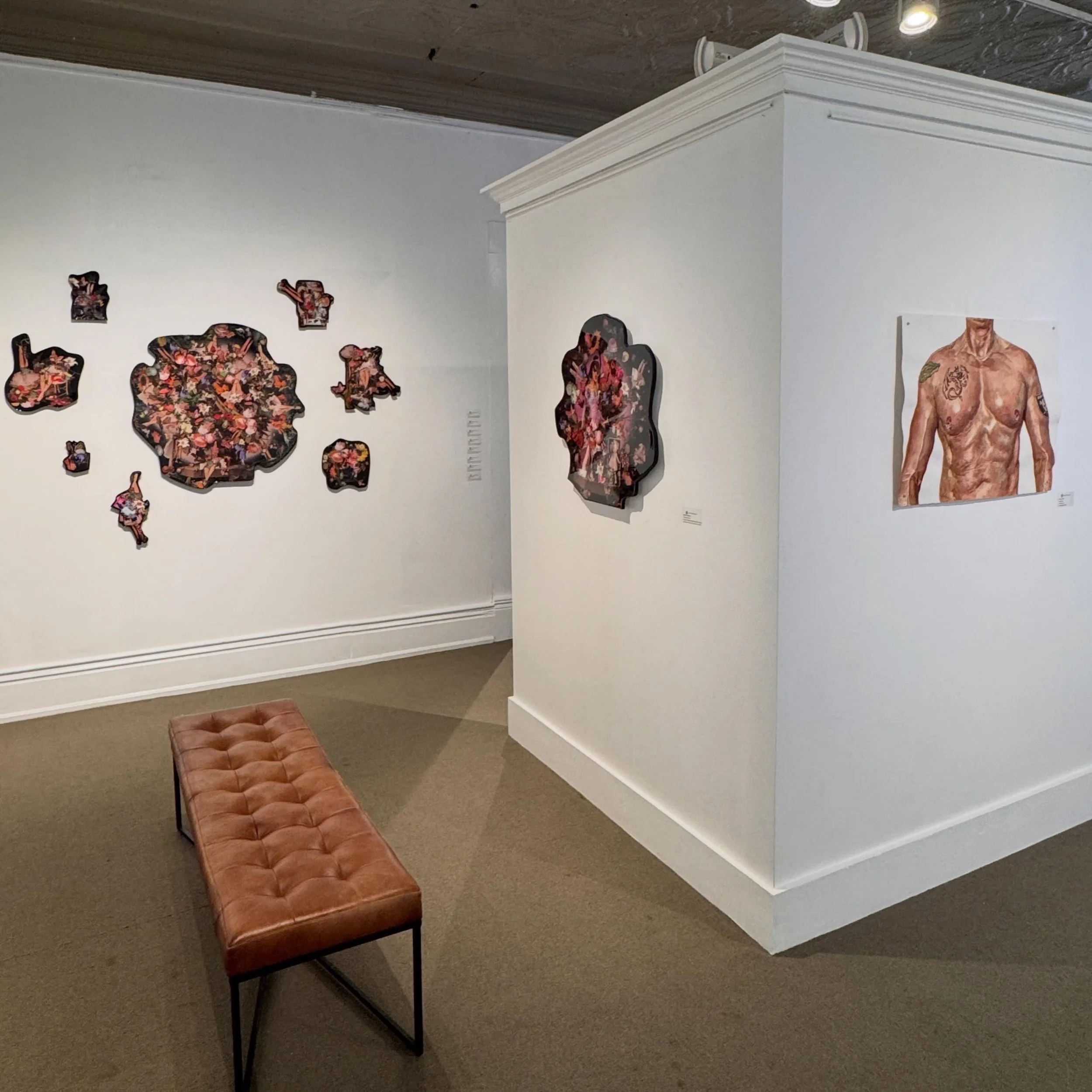 An art gallery with various paintings on the wall, a pink leather bench, and a white display case in the corner.