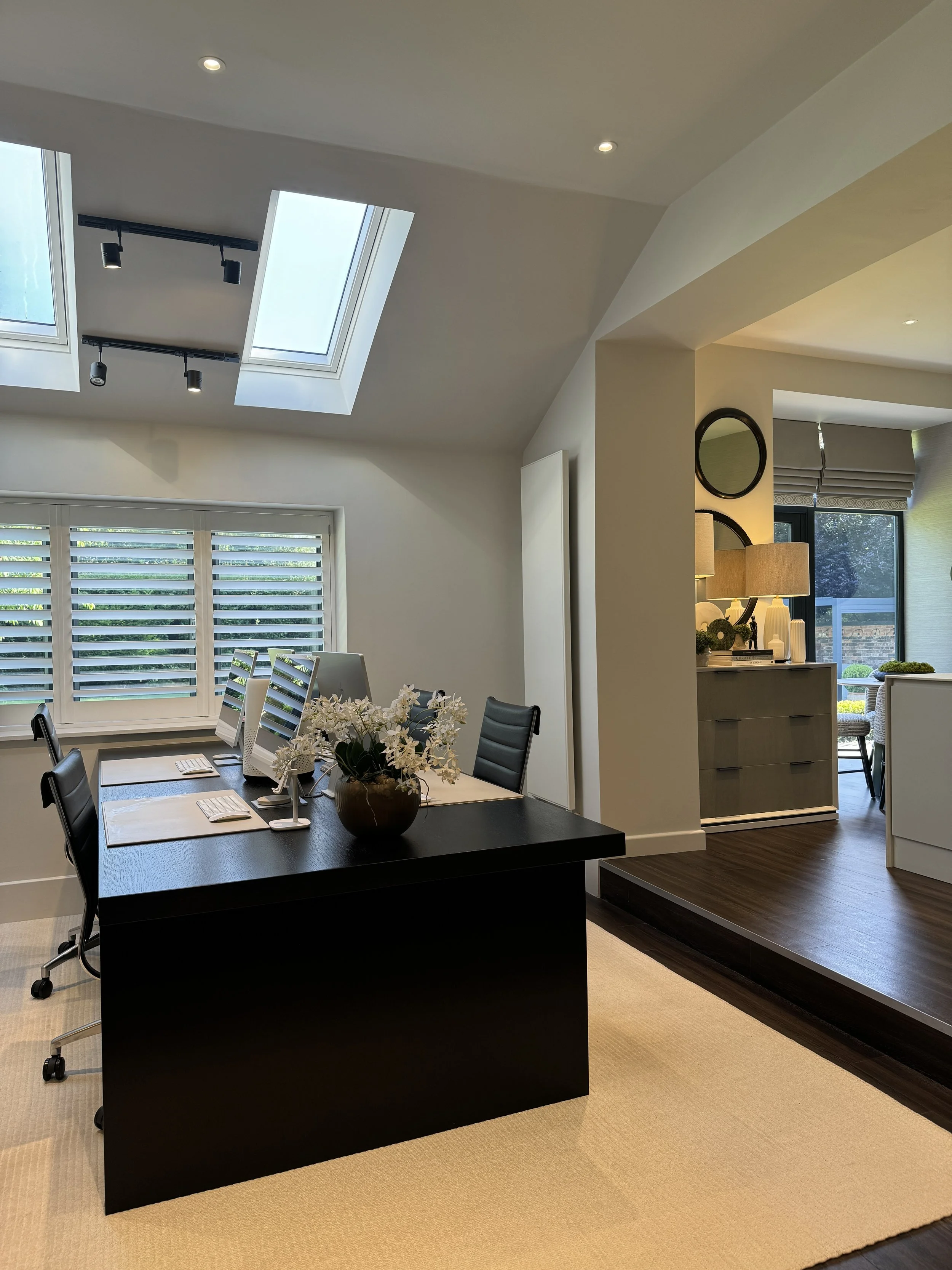Modern home office with a black desk, six computer monitors, chairs, and a beige rug, adjacent to a bright living area with lamps and large windows.