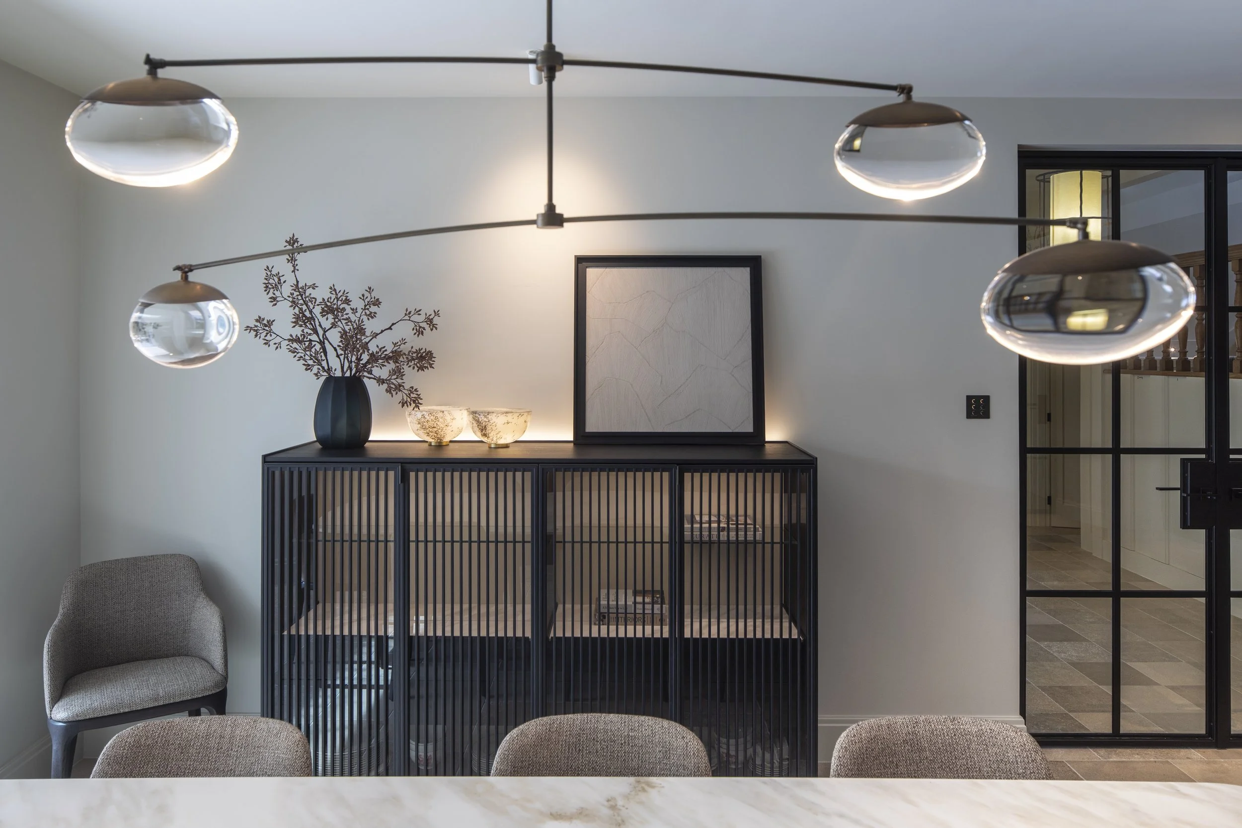 Interior of a modern living room with a stylish black console table, decorative bowls, a framed art piece, a vase with dried branches, a gray upholstered chair, and a large contemporary chandelier with glass globes hanging from a metal rod.