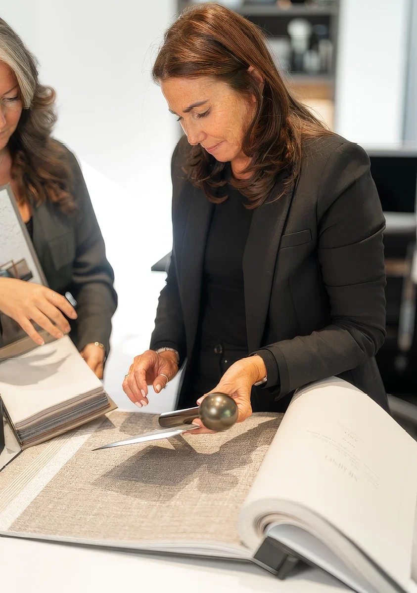 Two women examining color samples and design plans in an office, one is holding a phone and a color palette, the other is pointing at a large fabric swatch.