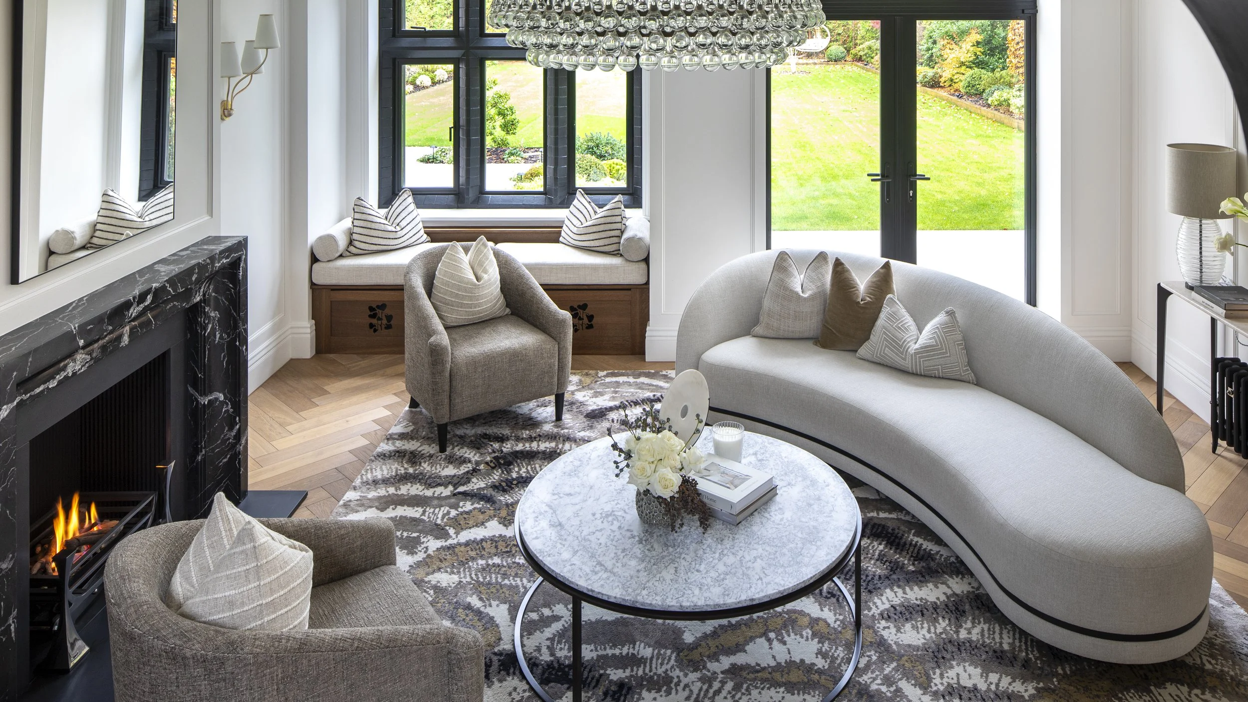 A living room with a curved white sofa, grey armchair, round marble coffee table, black marble fireplace, and large windows showing a green backyard.