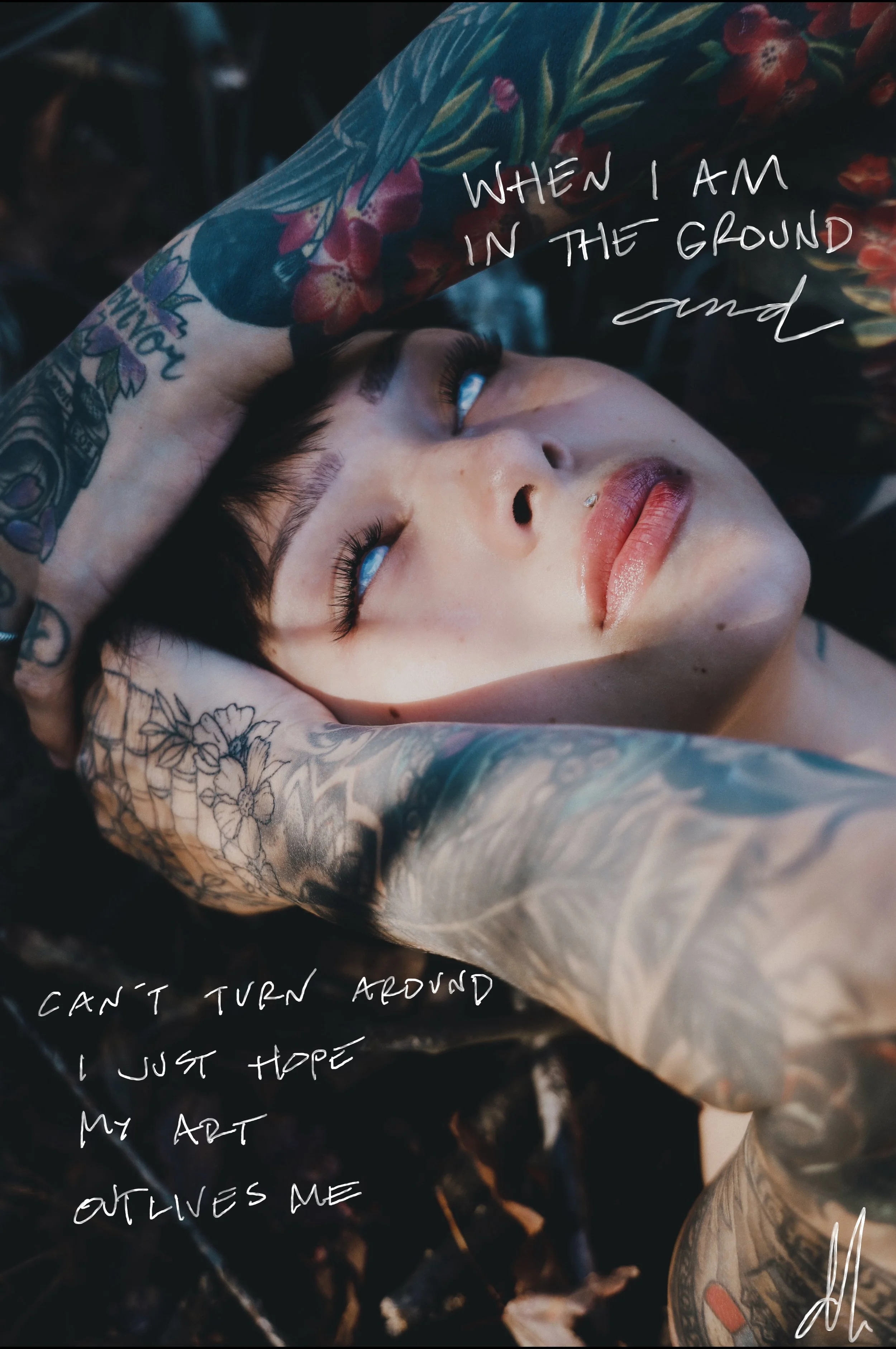 A woman with short dark hair and tattoos on her arms and face, lying outdoors with her hand on her head, gazing upward. Overlaid on the image are handwritten words: 'When I am in the ground and can't turn around I just hope my art outlives me.'