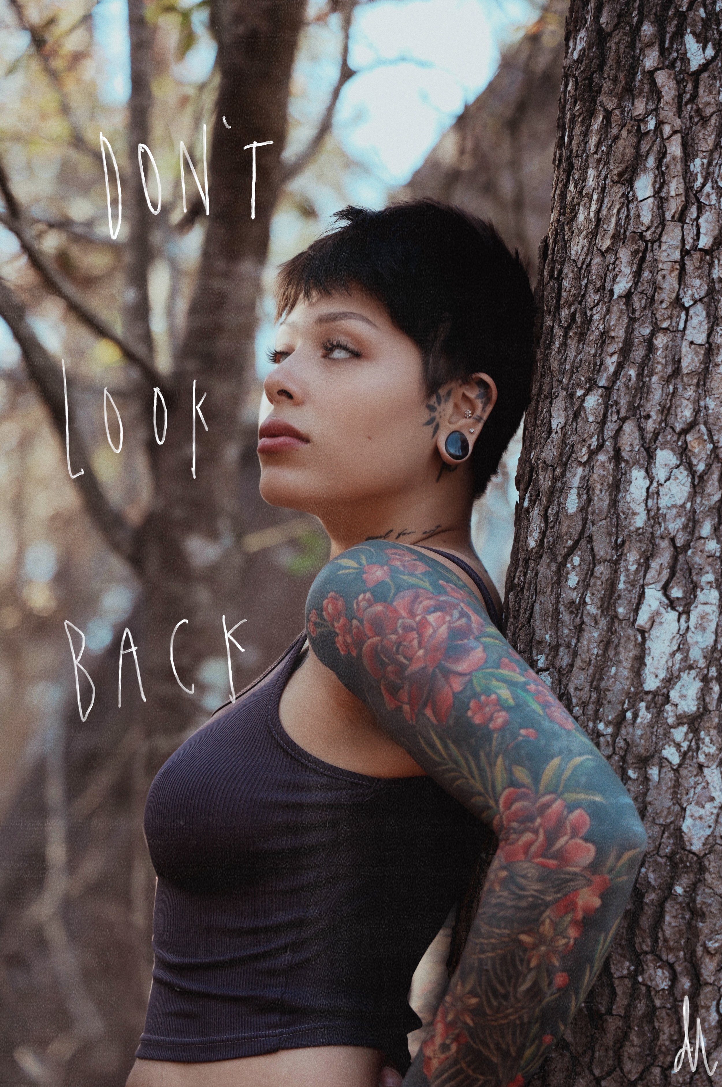 A woman with short dark hair and colorful tattoos on her arm, standing against a tree trunk in a forest. She is wearing a black tank top and looking away thoughtfully. The words 'Don't Look Back' are overlaid on the image.