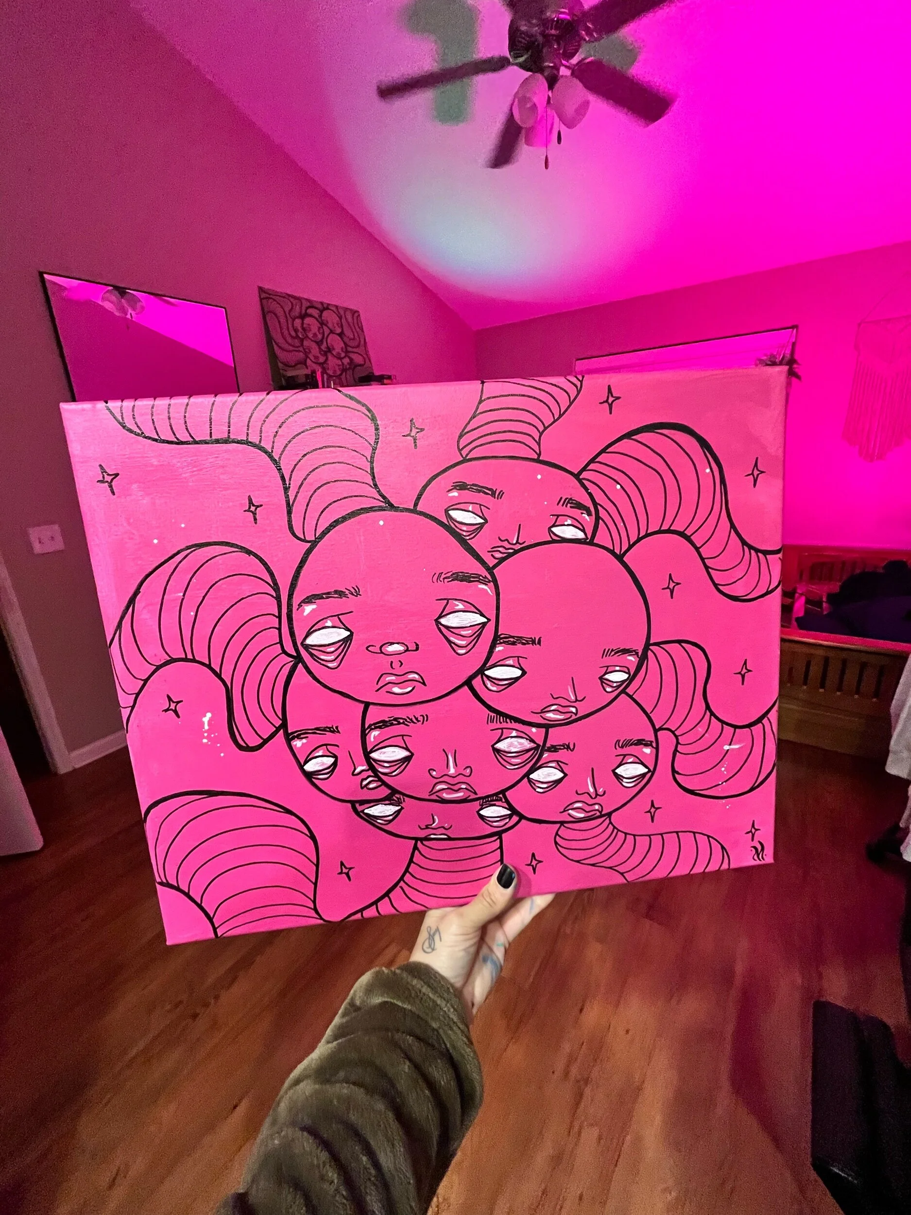 A person holding a pink illustration board with multiple faces and long hair, with a room illuminated in pink light in the background.