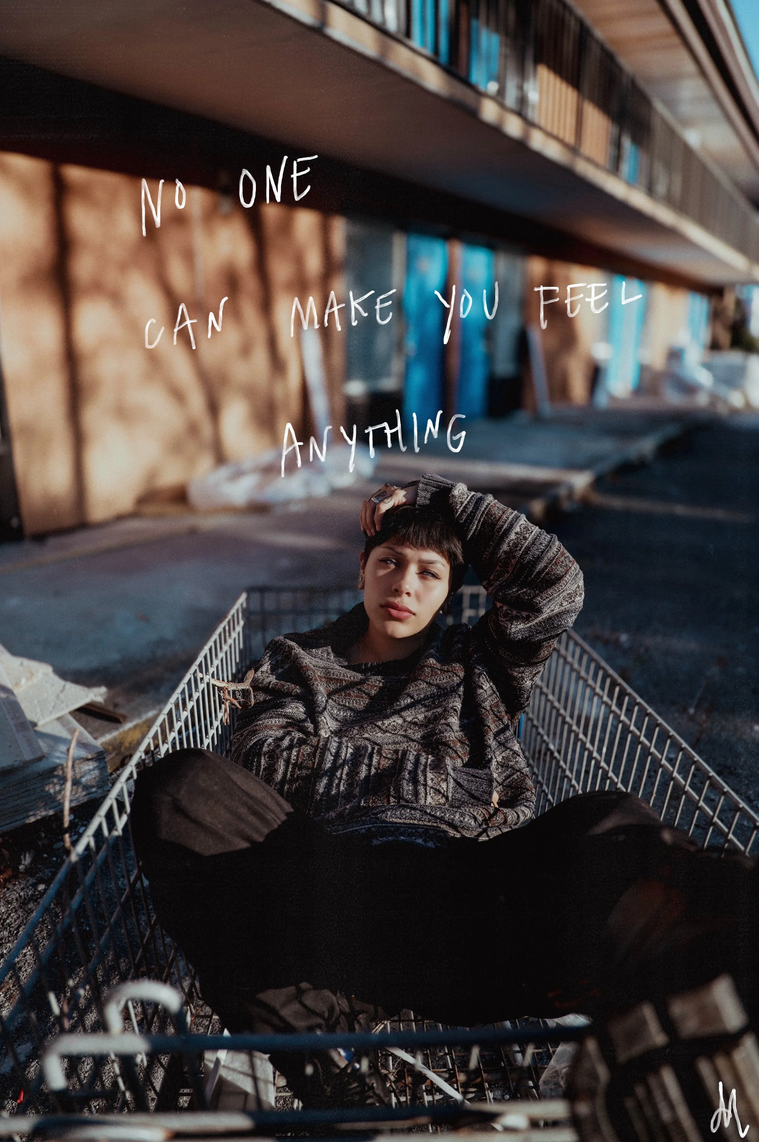 A young woman with short dark hair and casual clothing lying in a shopping cart outdoors, with a thoughtful expression, under a bridge or overpass with graffiti and bookshelves in the background, with the quote "No one can make you feel anything" wri