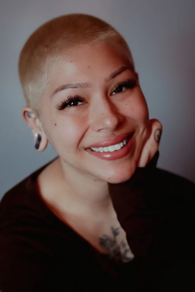 Young woman with short dark hair, glasses, and earrings in an art studio with paintings and art supplies in the background.