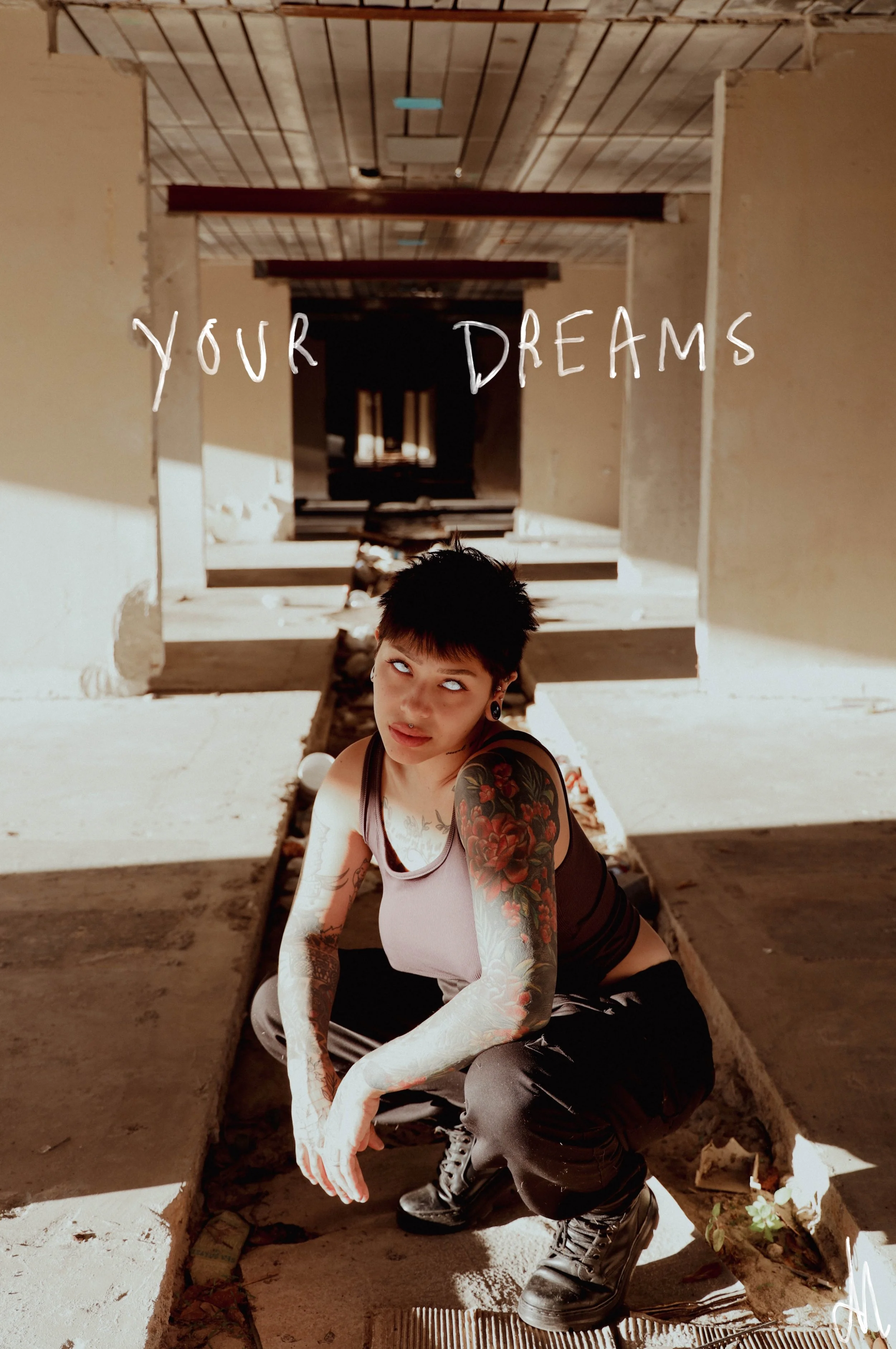 A person with short dark hair, tattoos, and piercings, wearing a tank top and black pants, crouching in an abandoned building with unfinished walls and flooring, with the phrase 'YOUR DREAMS' written across the top.
