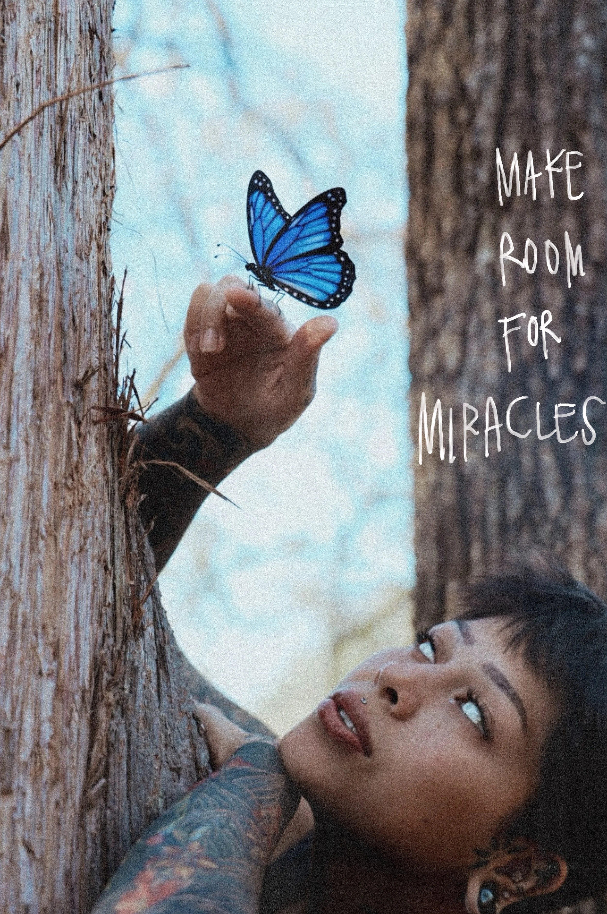 A person with short dark hair and tattoos on their arm, lying against a tree trunk, looking at a blue butterfly perched on their finger, with a background of trees and sky, and text that reads 'Make room for miracles'.