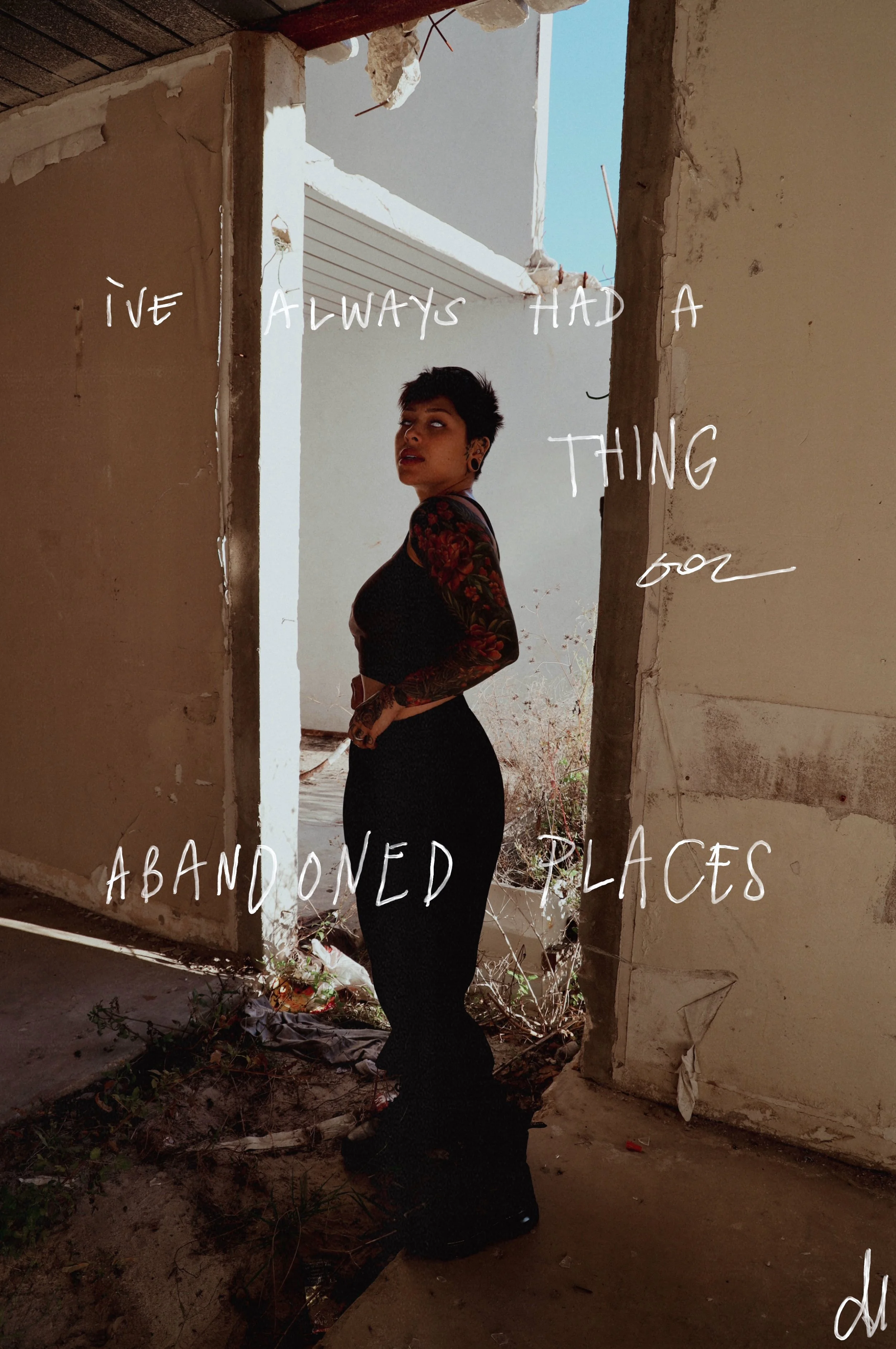 A person with short dark hair, tattoos on arms, wearing a black top and pants, standing in an abandoned, partially demolished building with overgrown weeds and debris on the ground. Text overlay reads, 'I've always had a thing for abandoned places.'